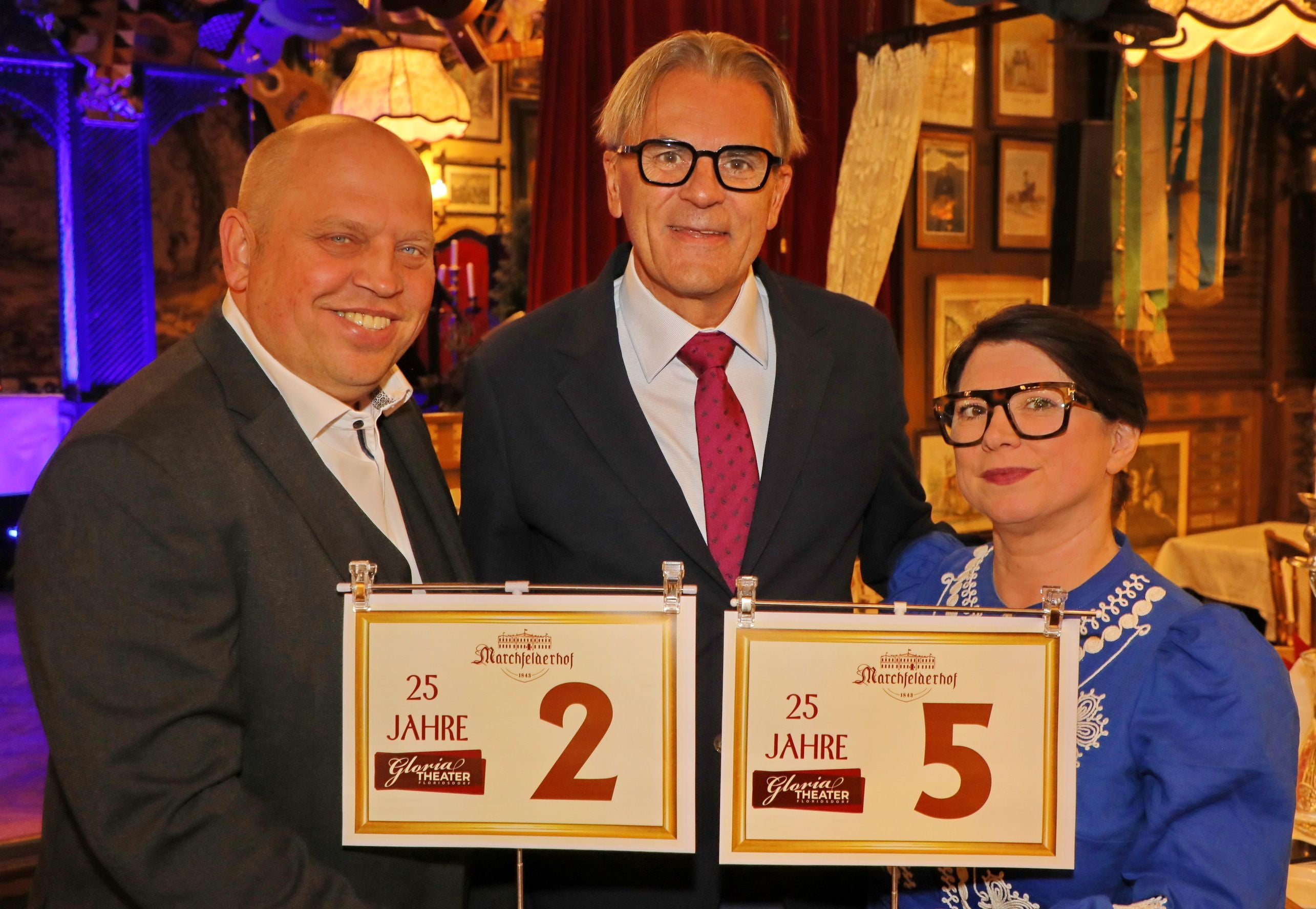 Drei Personen halten Schilder mit "25 Jahre Gloria Theater" und den Zahlen 2 und 5.