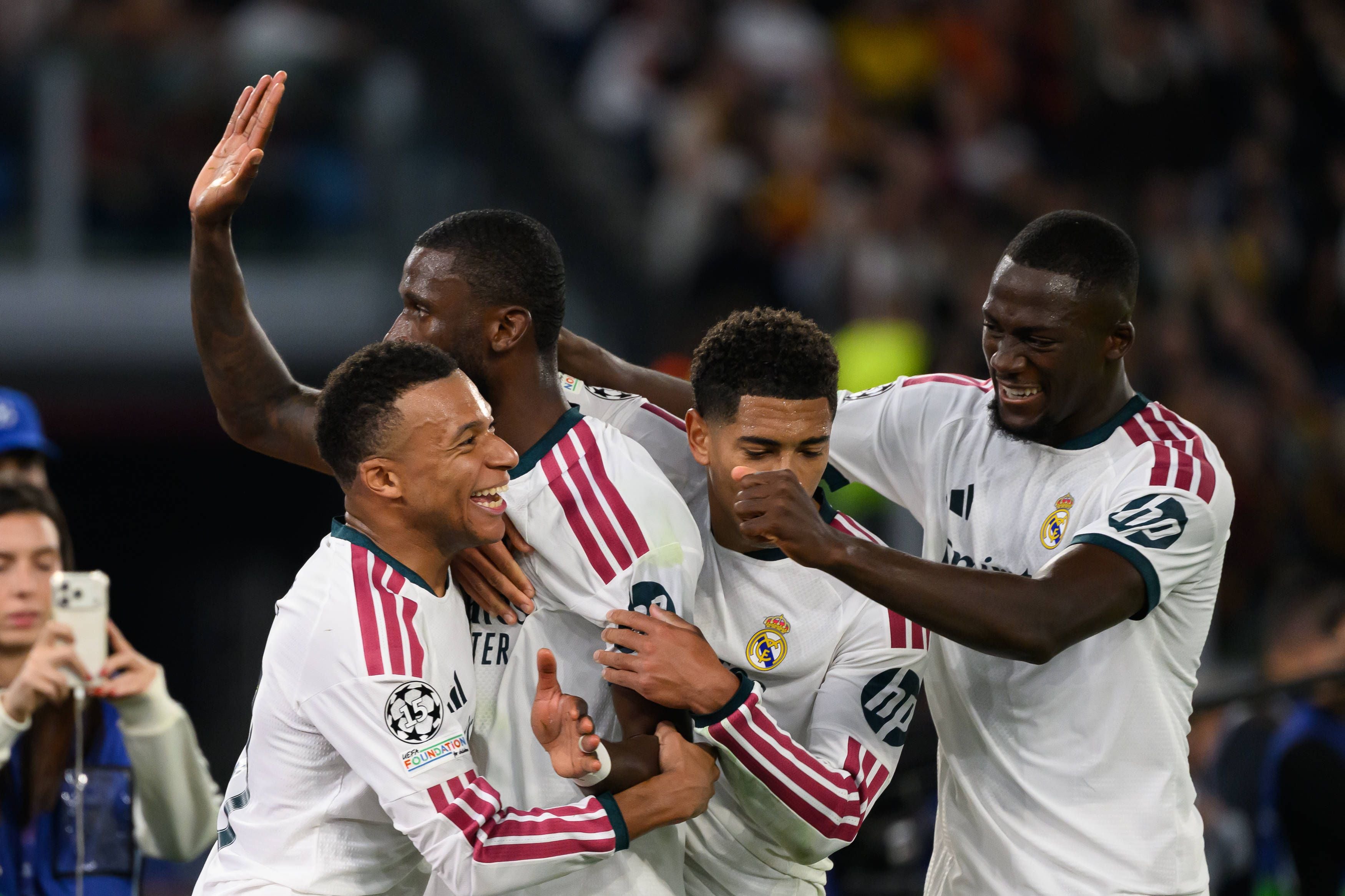 Kylian Mbappé, Jude Bellingham und Antonio Rüdiger jubeln gemeinsam mit weiteren Teamkollegen von Real Madrid im weiß-roten Auswärtstrikot herzhaft lachend auf dem Spielfeld.