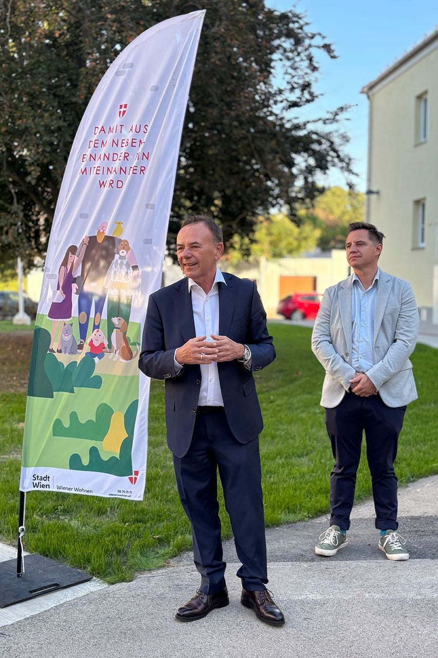 Zwei Männer in Anzügen stehen im Freien bei einer Veranstaltung vor einem Werbebanner.