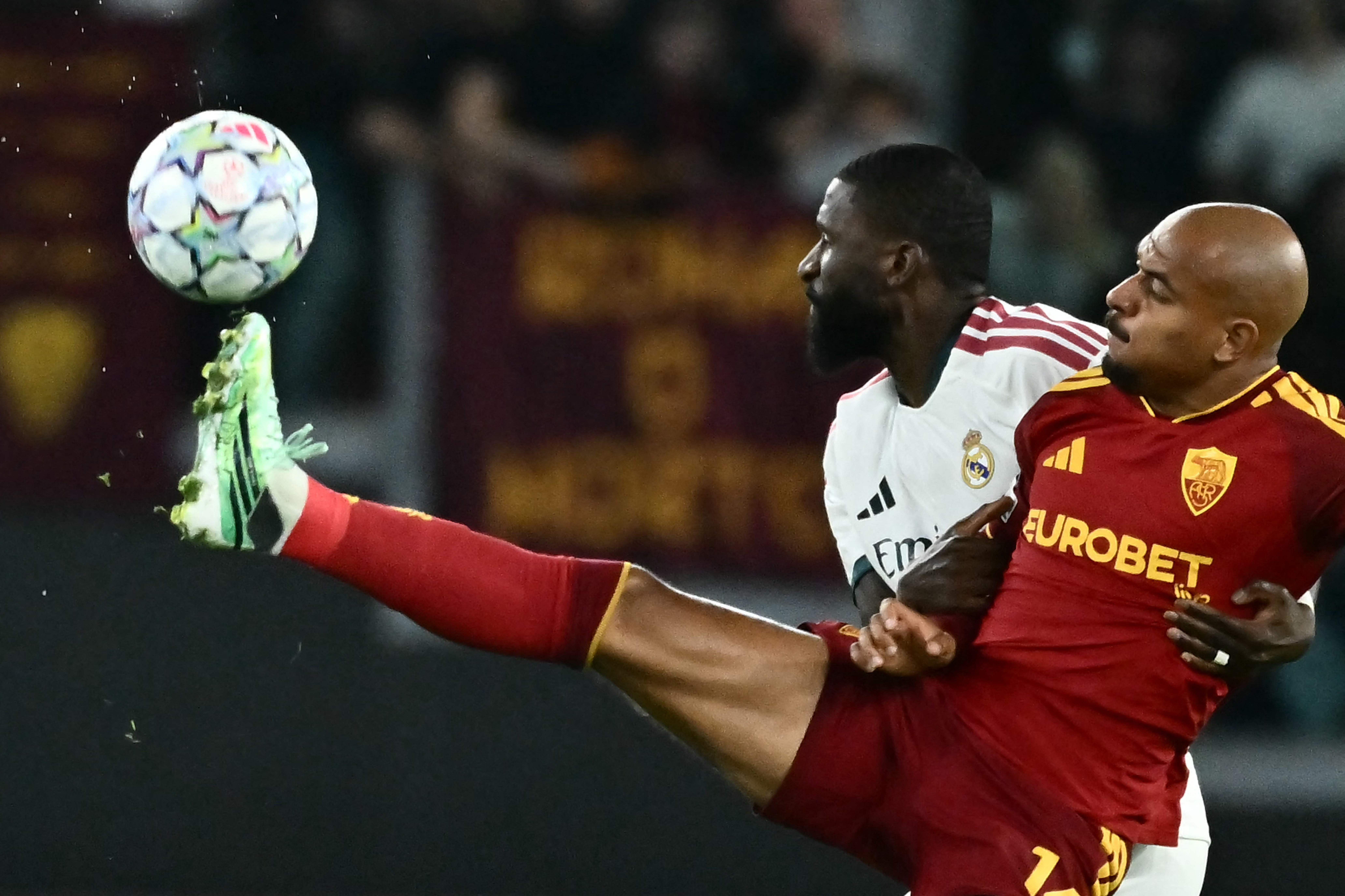 Antonio Rüdiger von Real Madrid setzt im weiß-roten Auswärtstrikot im Duell mit einem Roma-Gegenspieler im roten Dress zu einer akrobatischen Flugeinlage an, um den Ball mit dem Fuß aus der Luft zu spielen.