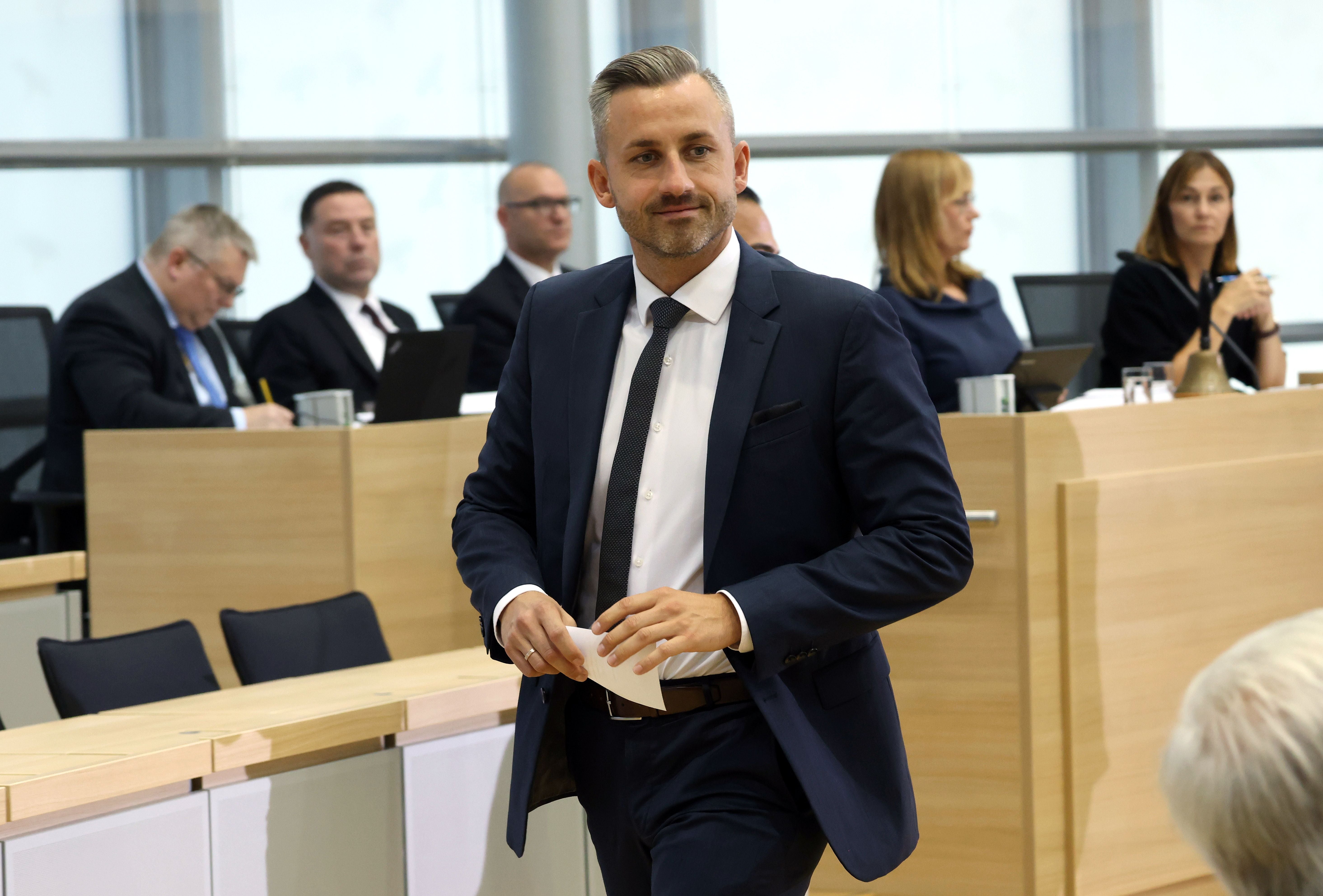 Ein Mann im Anzug geht durch einen modernen Sitzungssaal mit mehreren sitzenden Personen im Hintergrund.