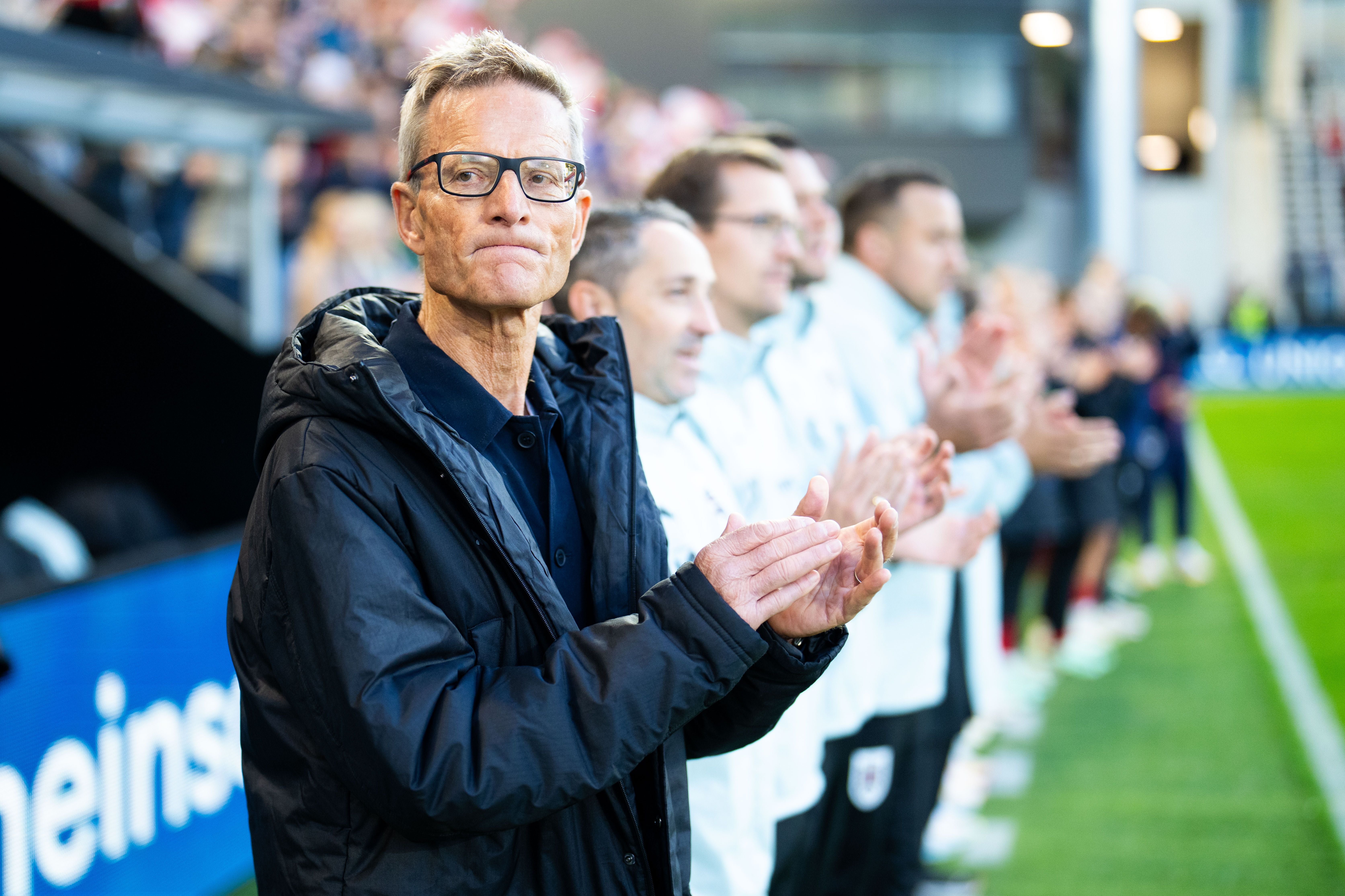 Ein Mann im schwarzen Mantel steht am Spielfeldrand und klatscht, weitere Personen im Hintergrund.