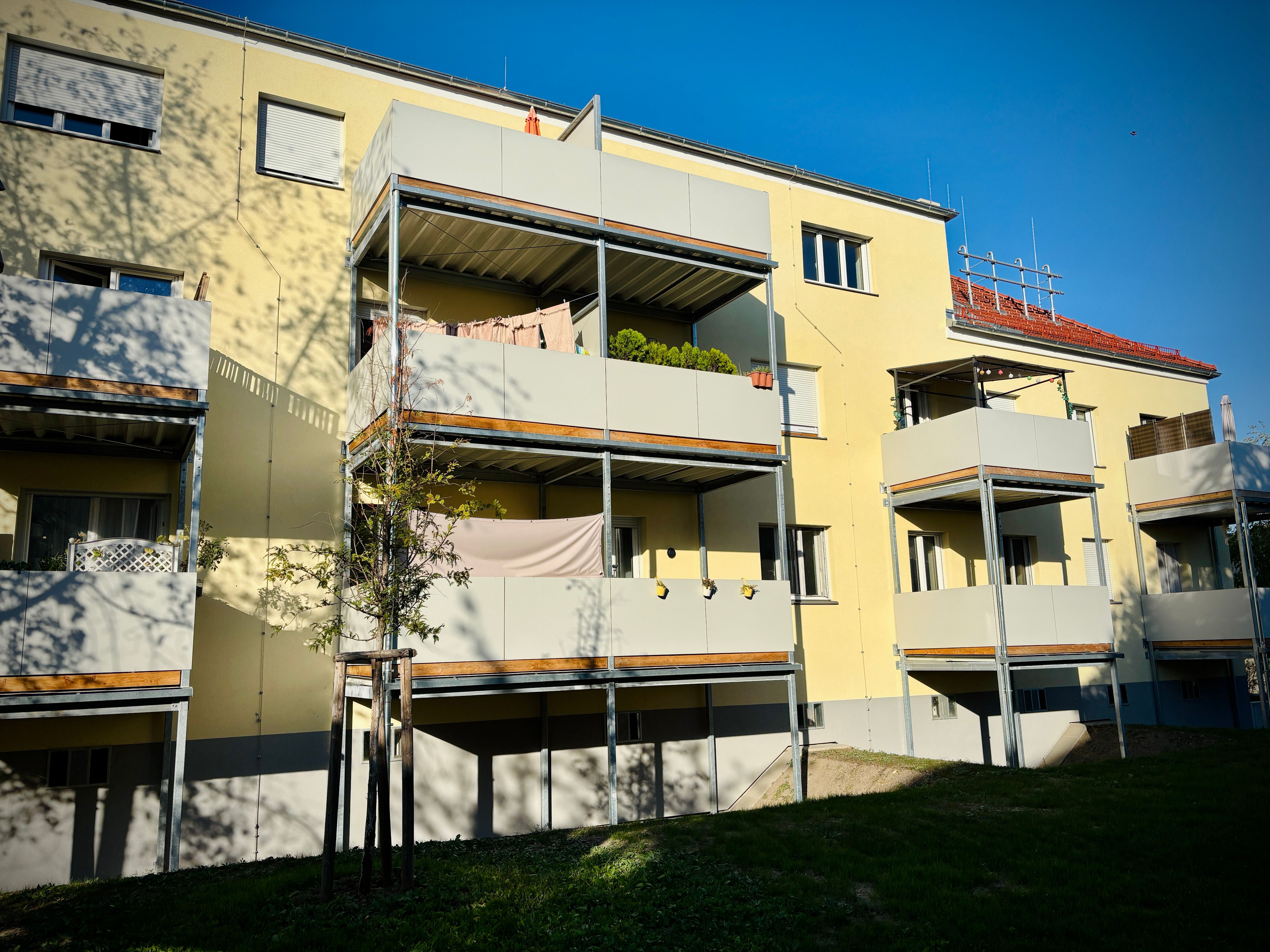 Mehrfamilienhaus mit Balkonen, gelber Fassade und Schatten von Bäumen im Vordergrund.