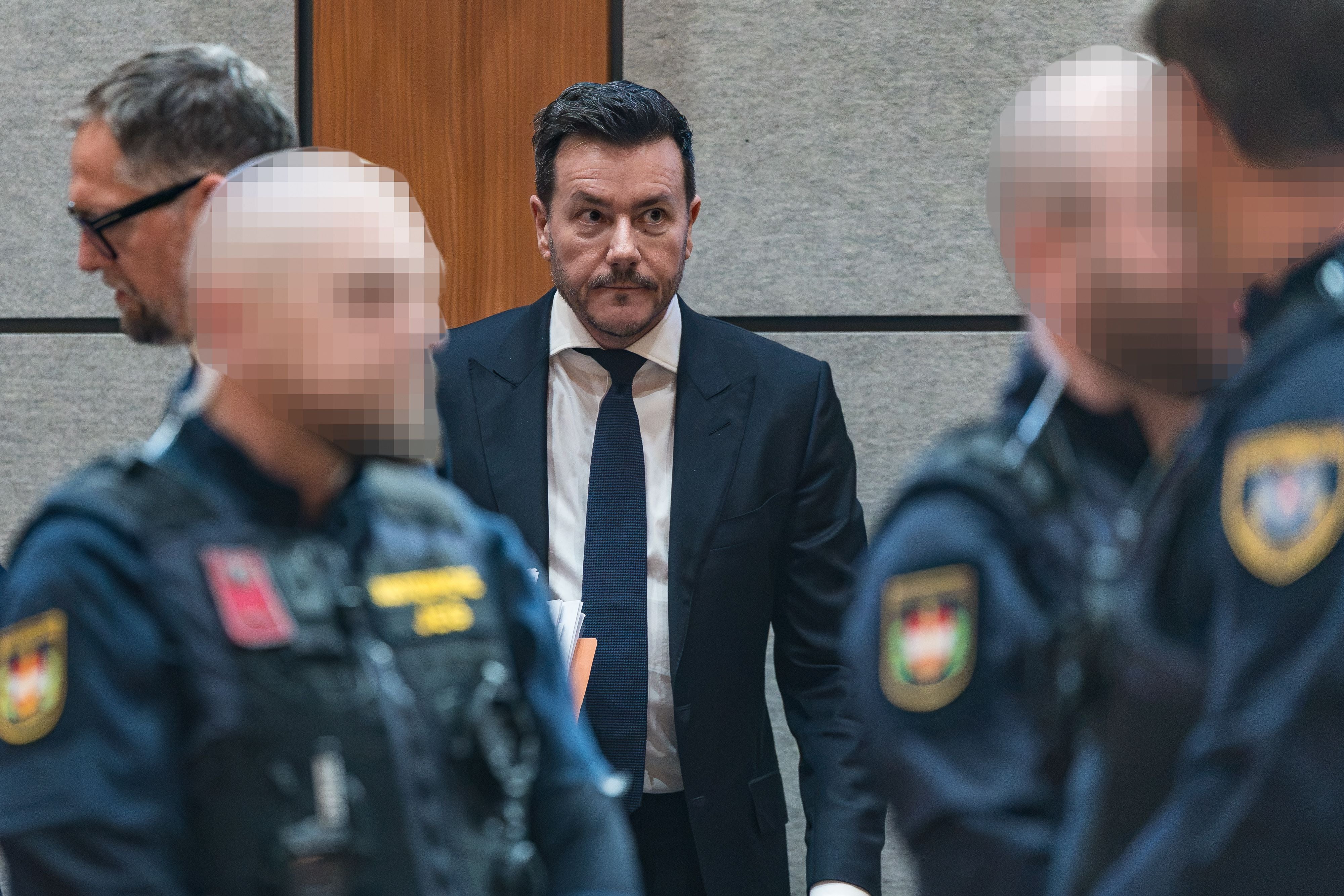 Ein Mann im Anzug steht in einem Gerichtssaal, umgeben von Polizisten mit unscharfen Gesichtern.