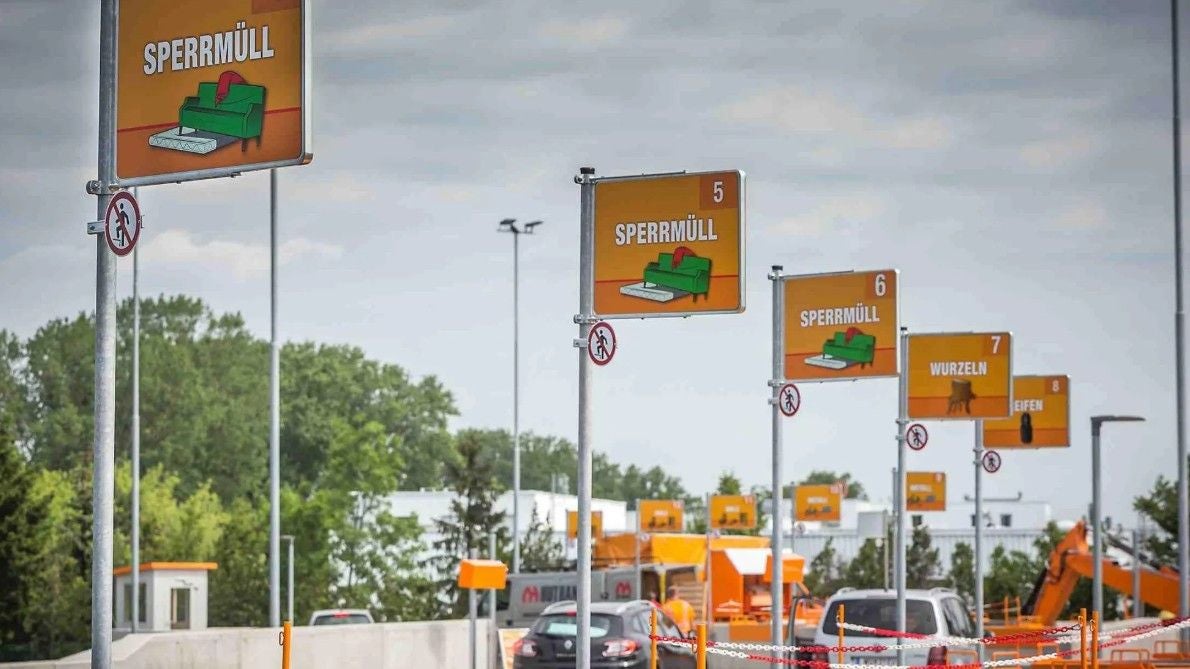 Autos und Schilder für Mülltrennung auf dem Mistplatz Ottakring bei bewölktem Himmel.