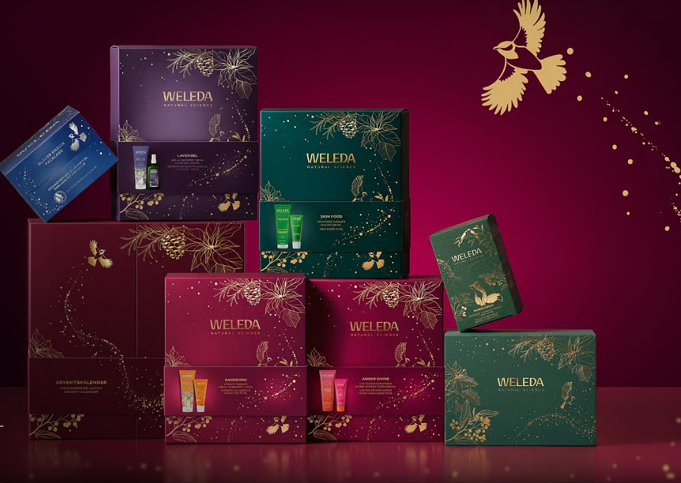 Verschiedene festliche WELEDA-Geschenkboxen vor bordeauxrotem Hintergrund mit goldenen Motiven.