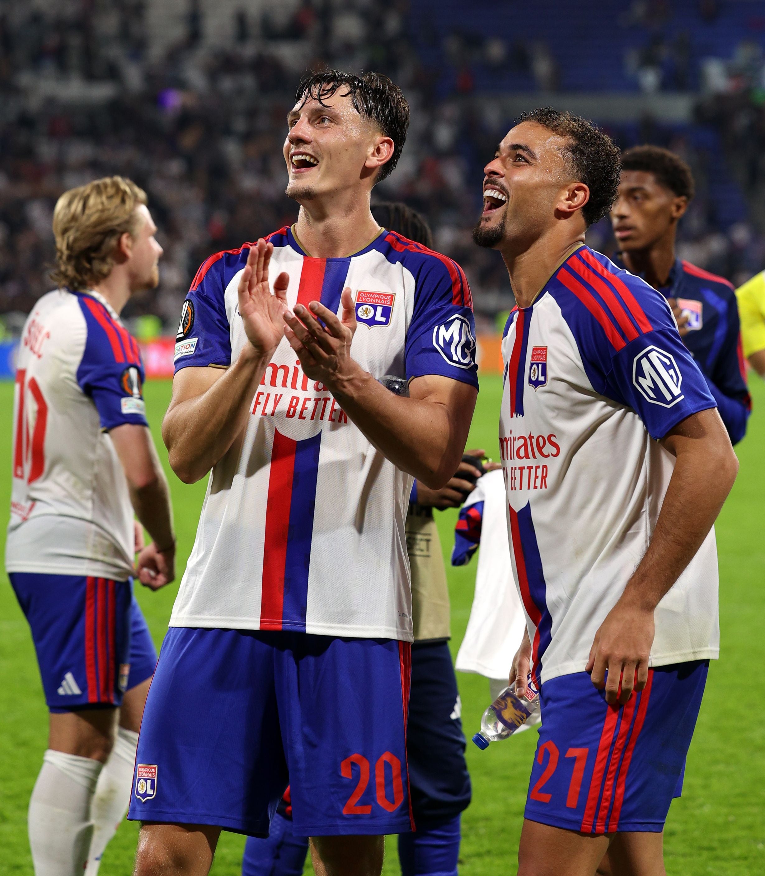 Felix Bacher und ein Teamkollege von Olympique Lyon jubeln strahlend im weiß-blau-roten Trikot mit erhobenen Händen vor den Fans auf dem Spielfeld.