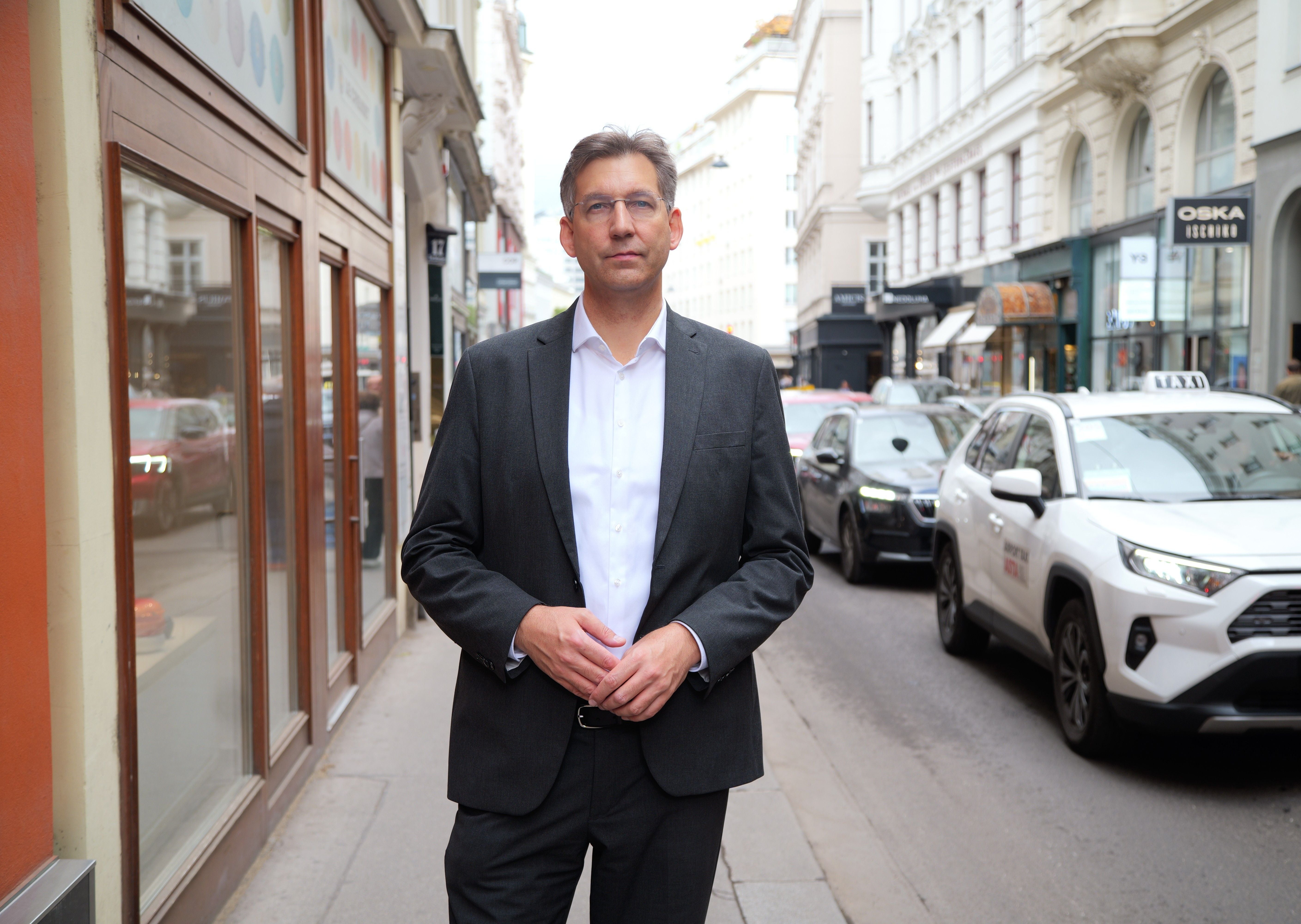 Ein Mann im Anzug steht auf einem Gehweg in einer belebten Innenstadt mit Autos im Hintergrund.
