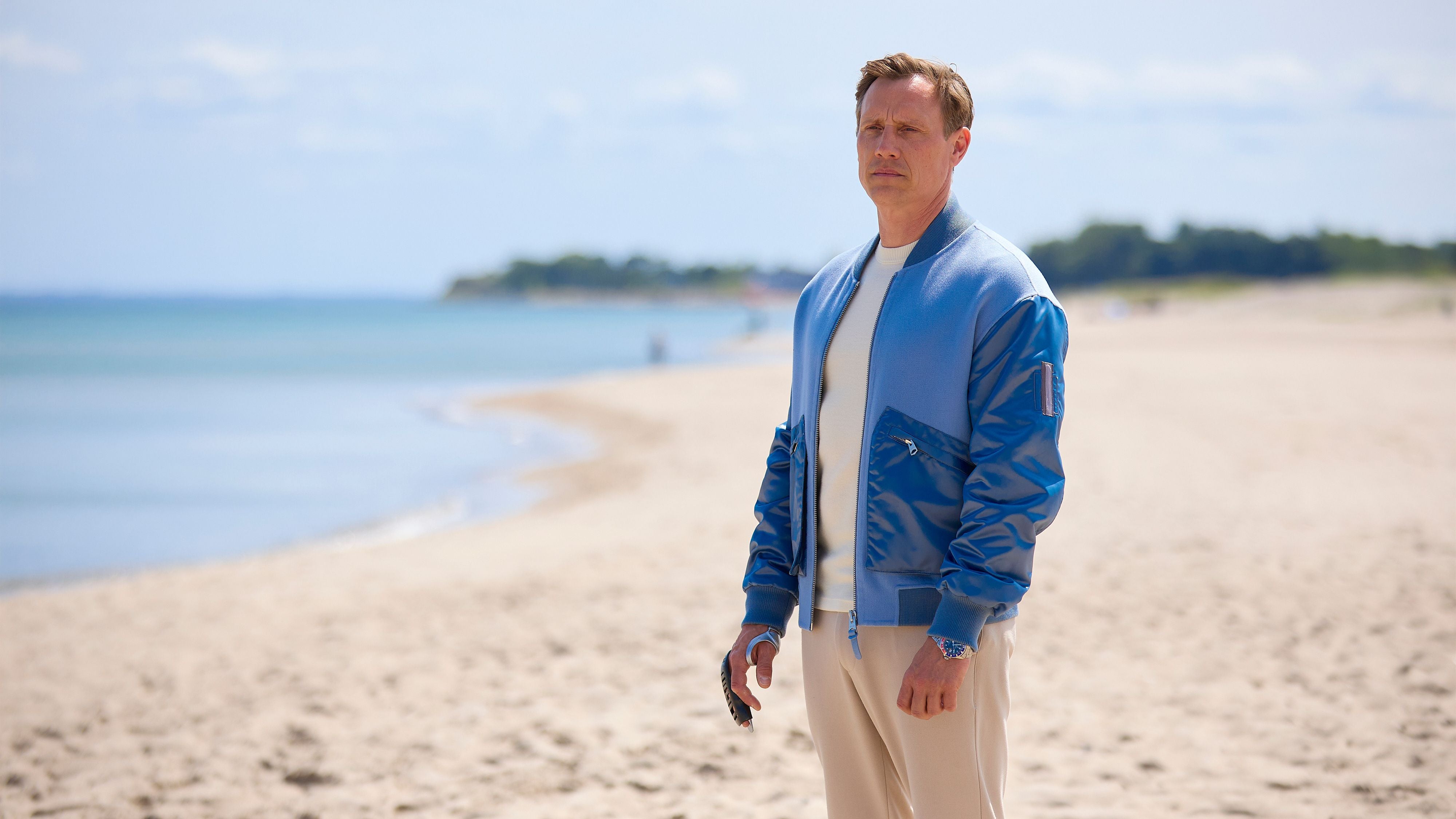 Ein Mann im blauen Blouson steht nachdenklich am Sandstrand mit Blick aufs Meer.
