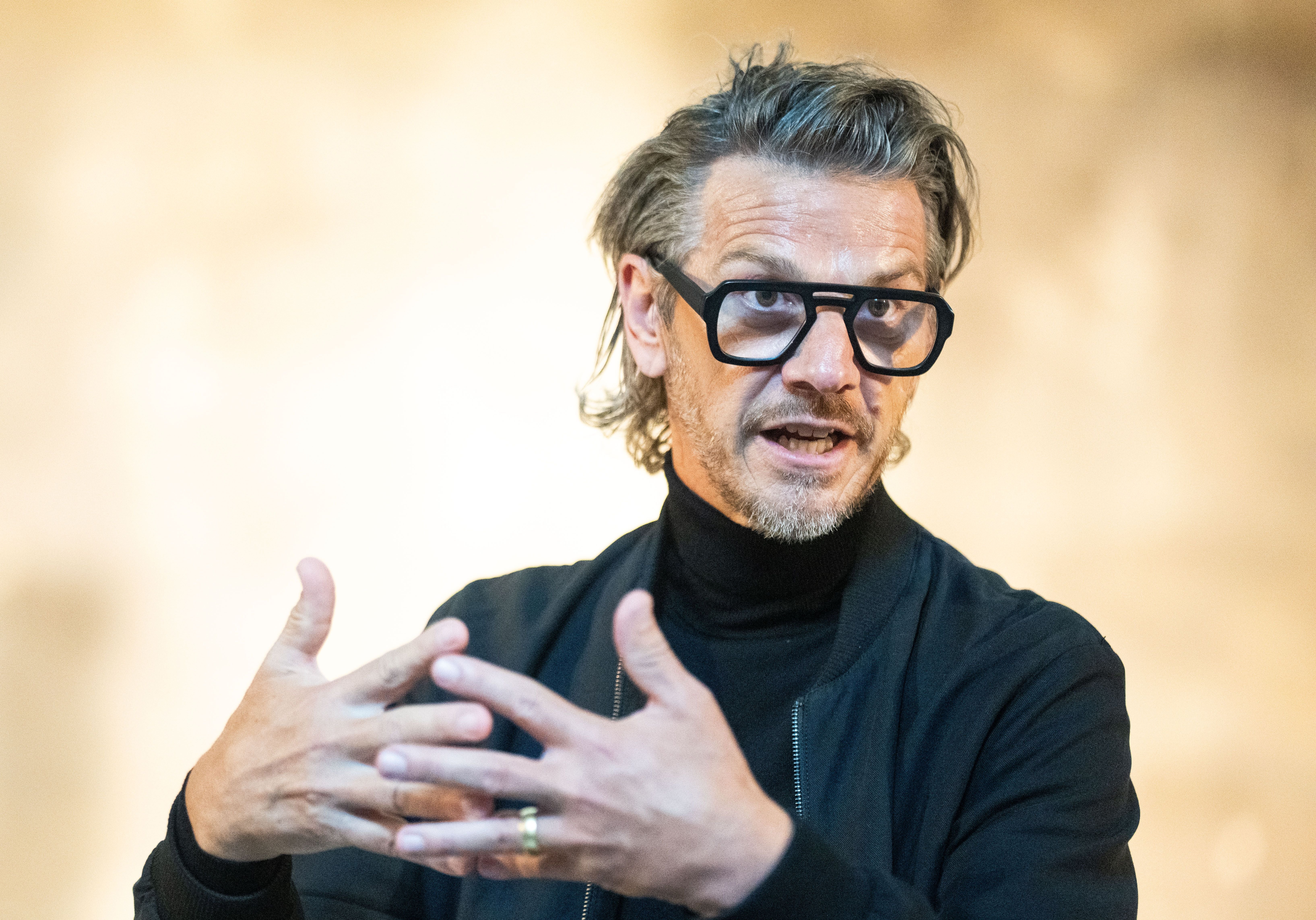 Ein Mann mit Brille und Bart spricht gestikulierend in einem Gespräch vor unscharfem Hintergrund.
