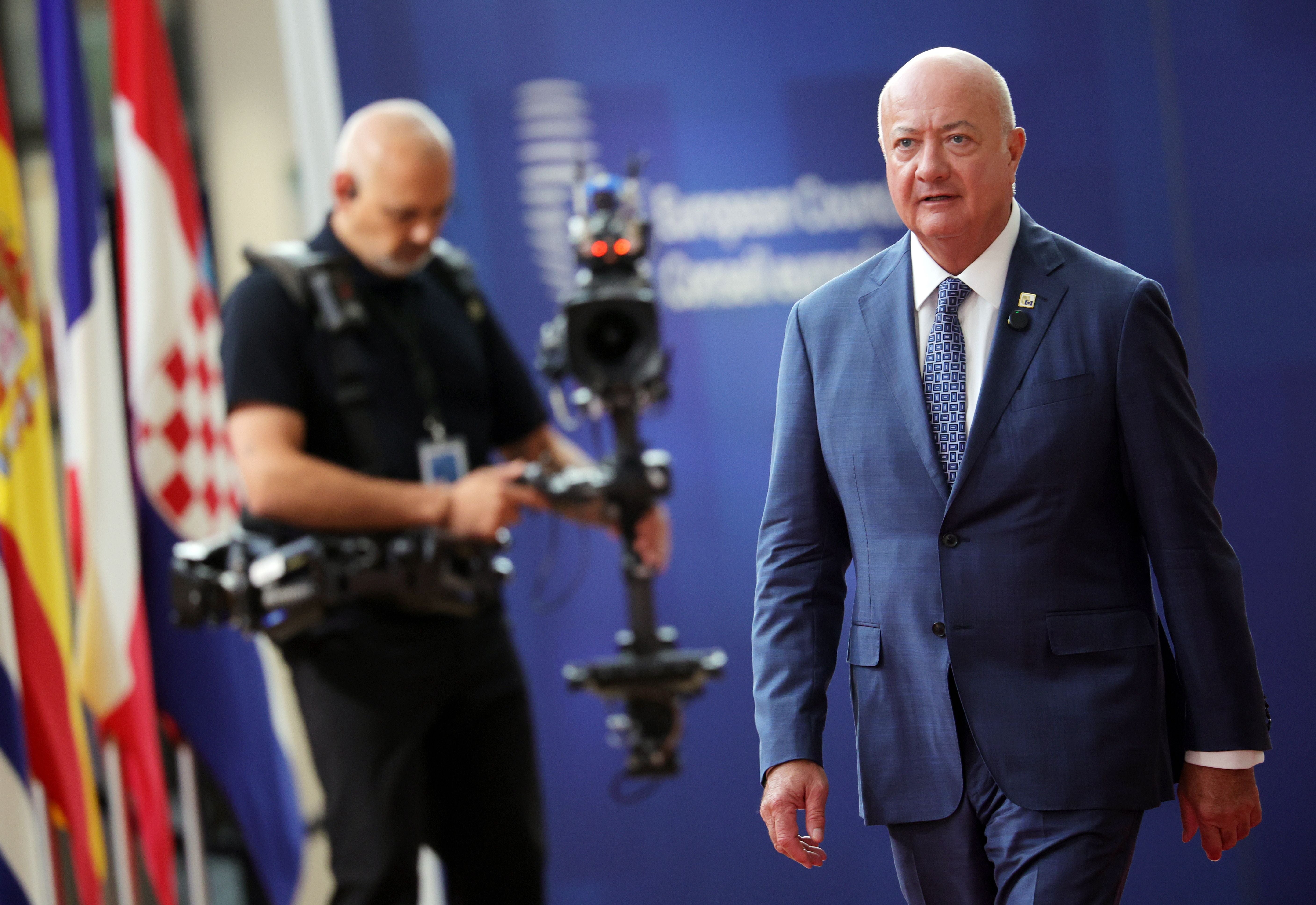 Ein Mann im blauen Anzug geht, im Hintergrund Kameramann und verschiedene EU-Flaggen.