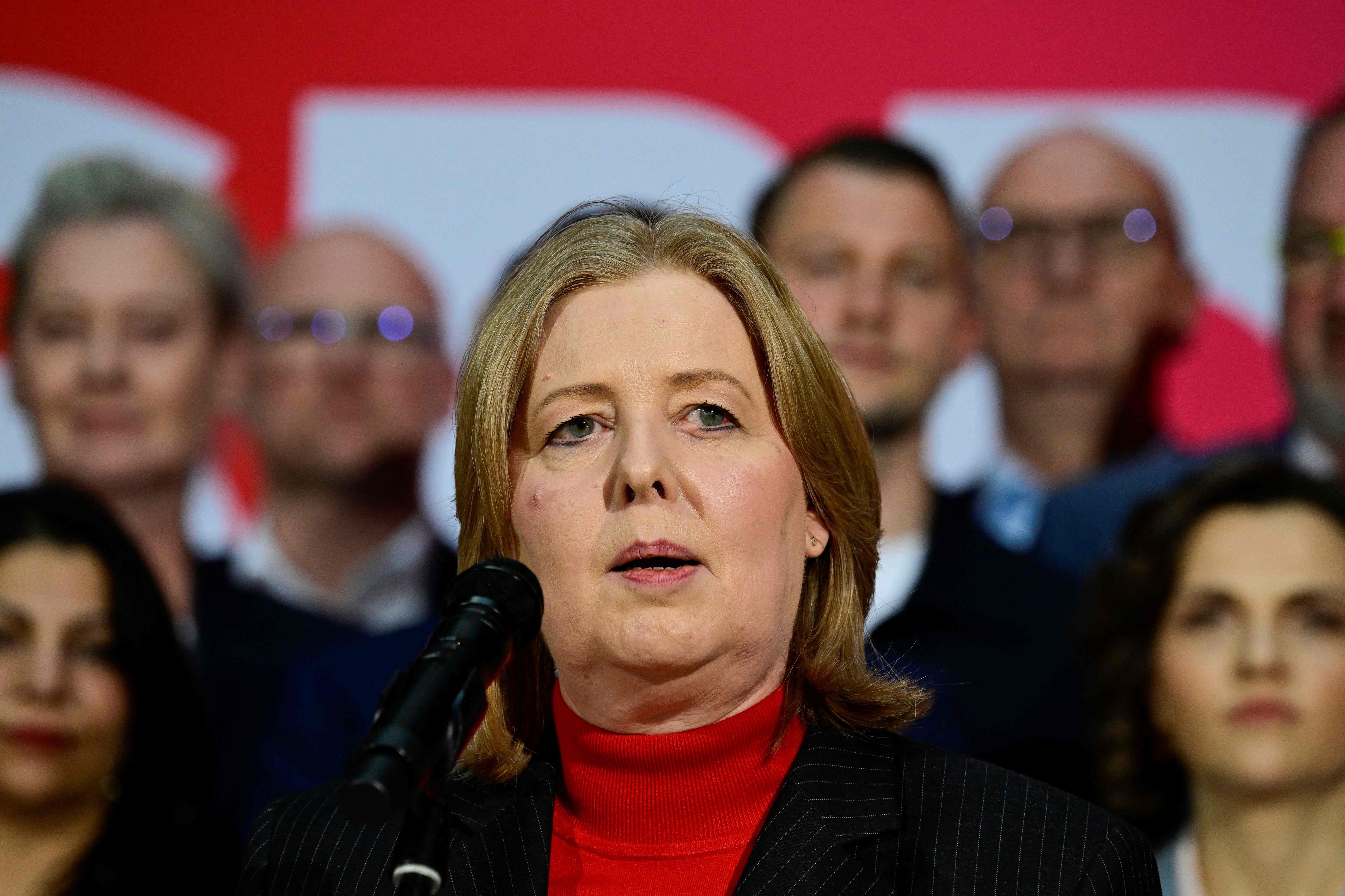 Eine Frau spricht am Mikrofon, im Hintergrund stehen mehrere Personen unscharf.