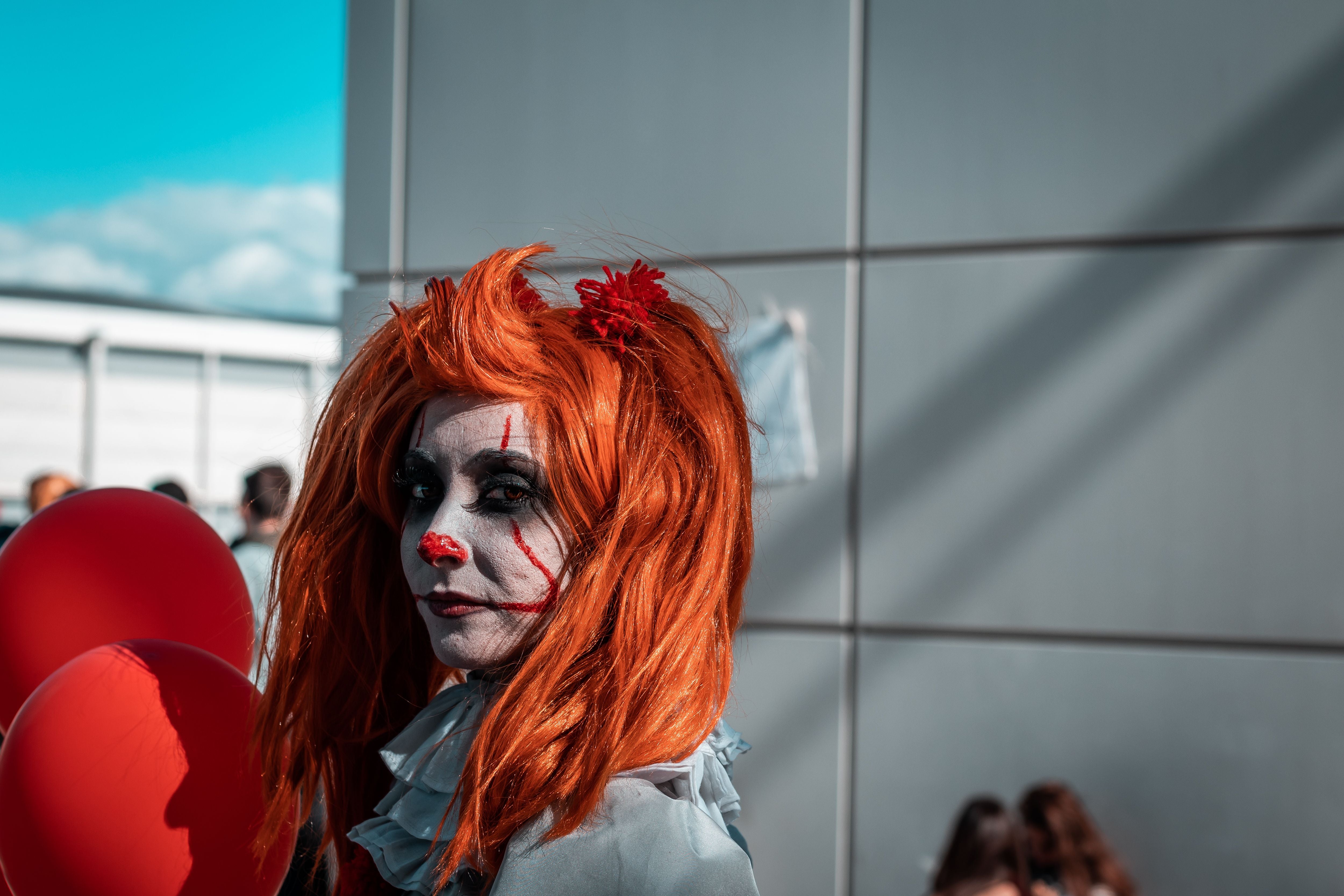 Junge Frau als Pennywise mit roter Perücke und Clown-Make-up, hält rote Ballons auf einer Messe.