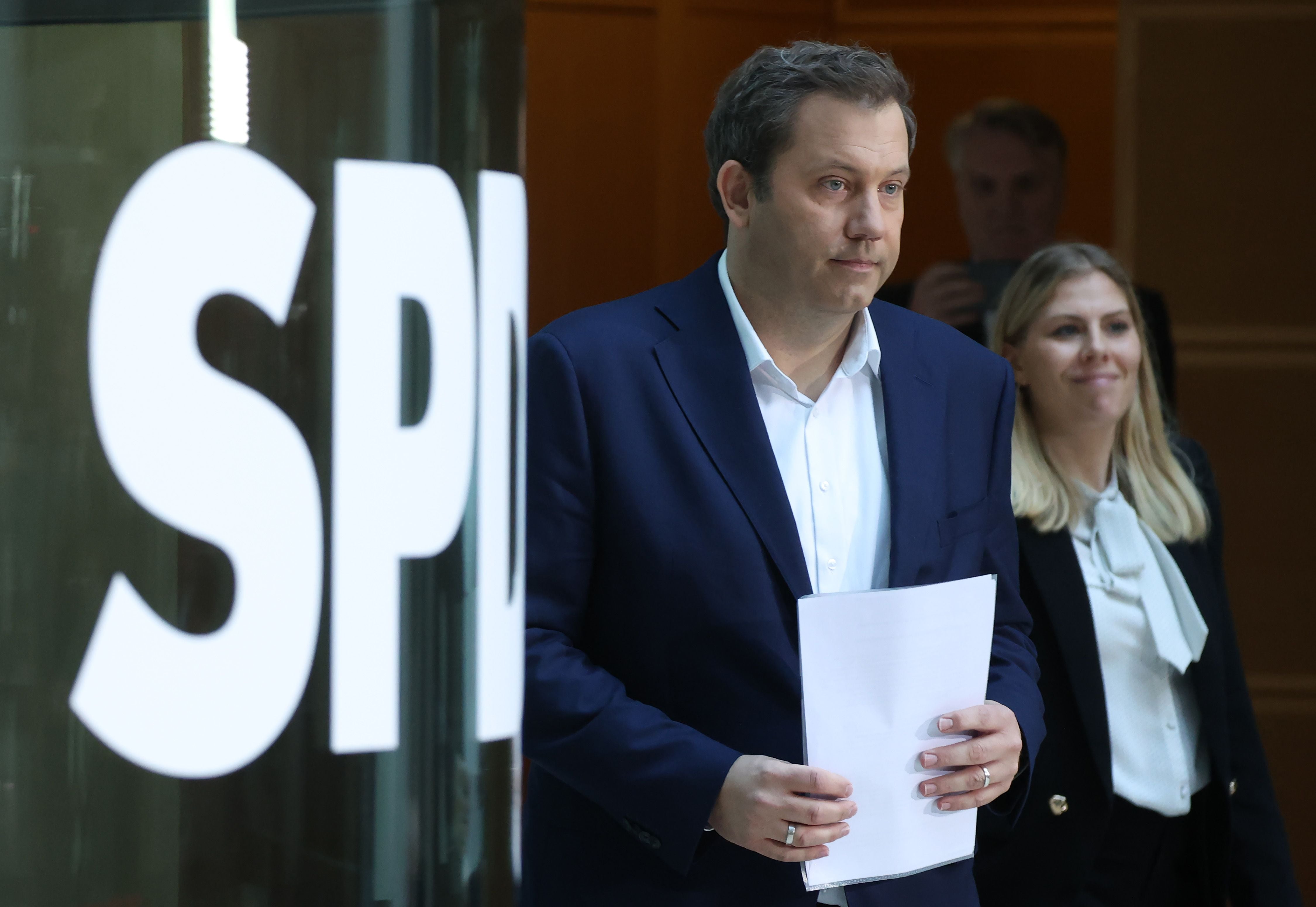 Lars Klingbeil steht mit Dokumenten vor einem SPD-Logo, eine Frau folgt ihm im Hintergrund.