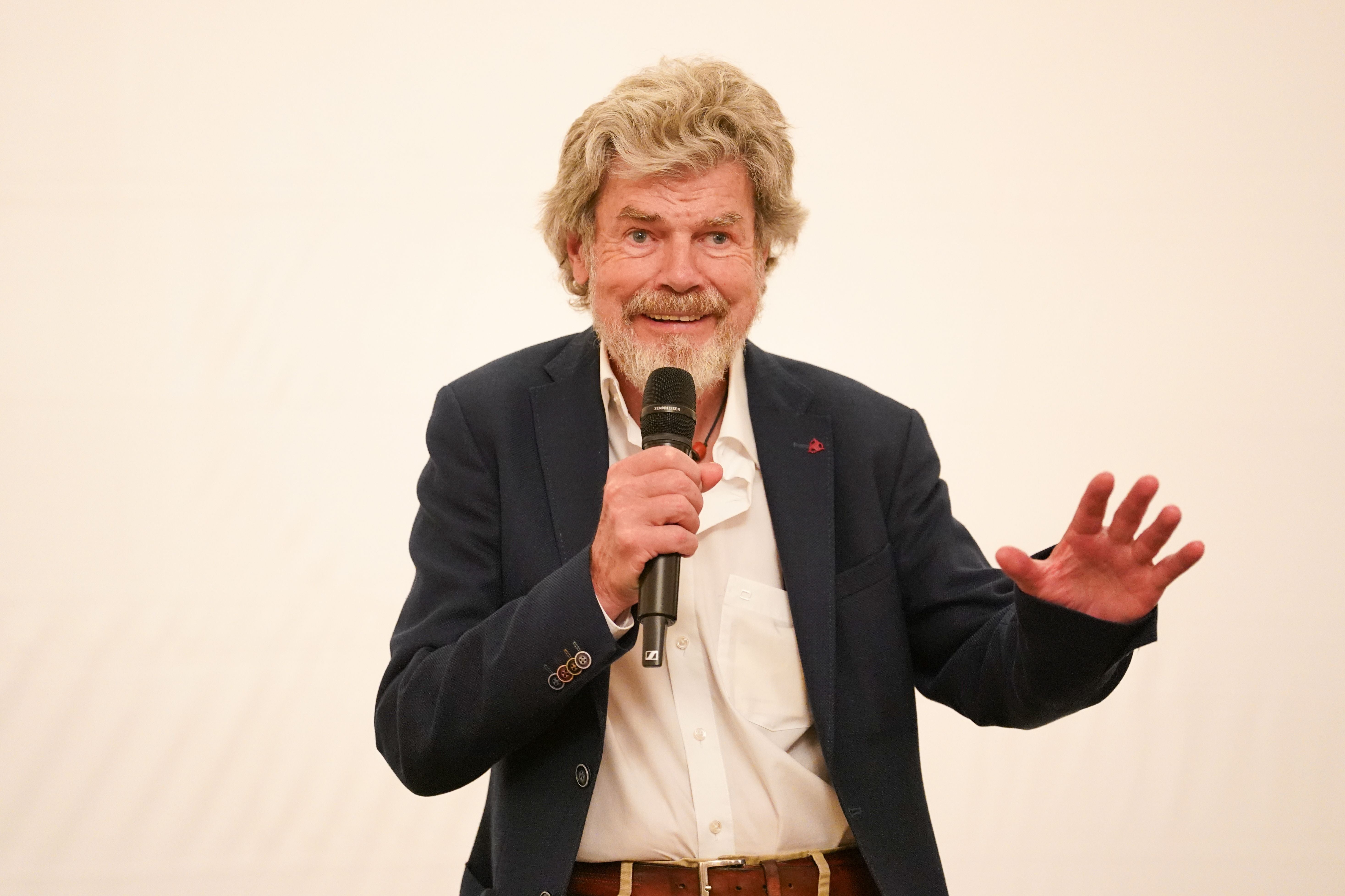 Ein älterer Mann mit Mikrofon spricht auf einer Bühne vor hellem Hintergrund.