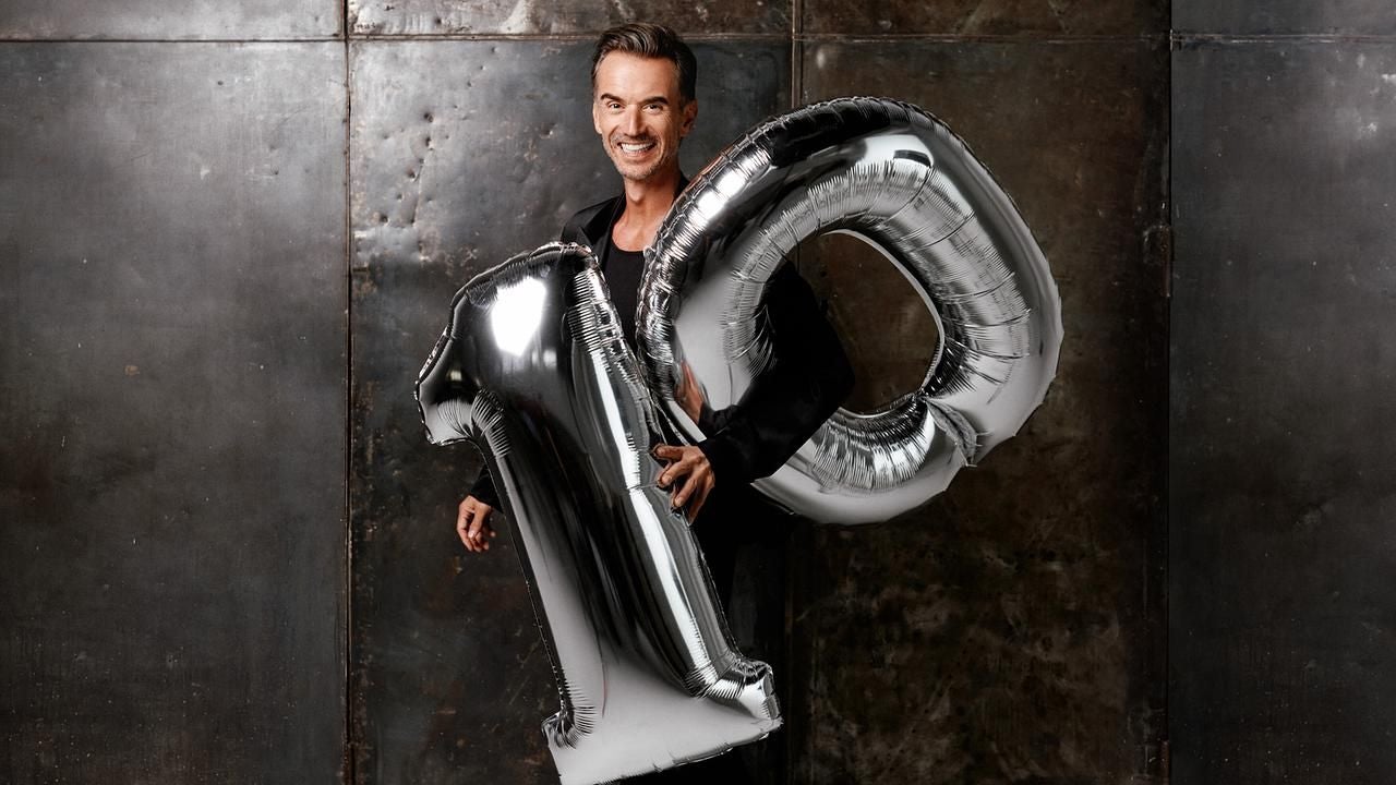 Mann hält große silberne Luftballons in Form der Zahl 10 vor dunklem Hintergrund.