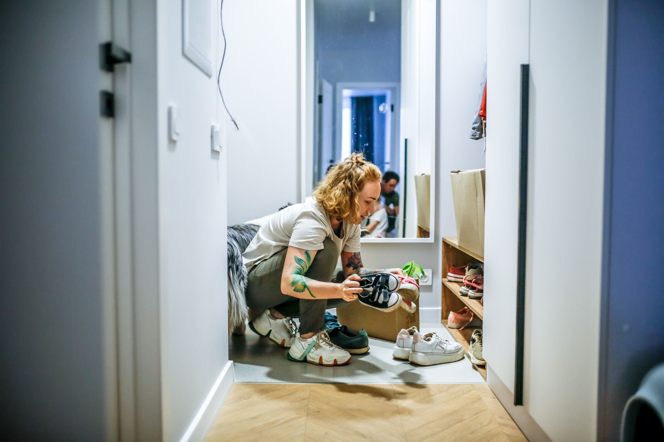 Frau mit Tattoos sortiert im Flur zuhause Schuhe vor einem Regal.