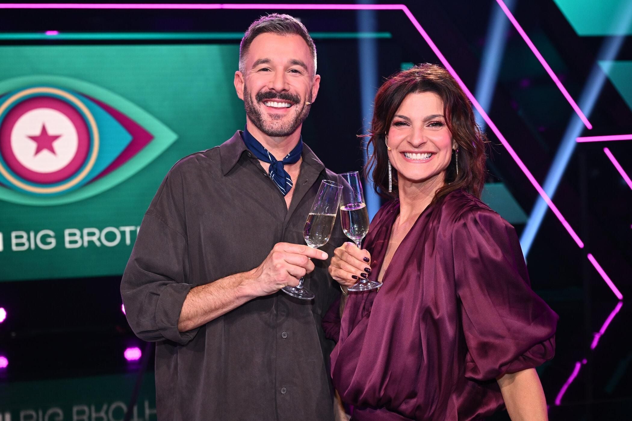 Zwei lächelnde Personen stoßen mit Sektgläsern in einem TV-Studio an.