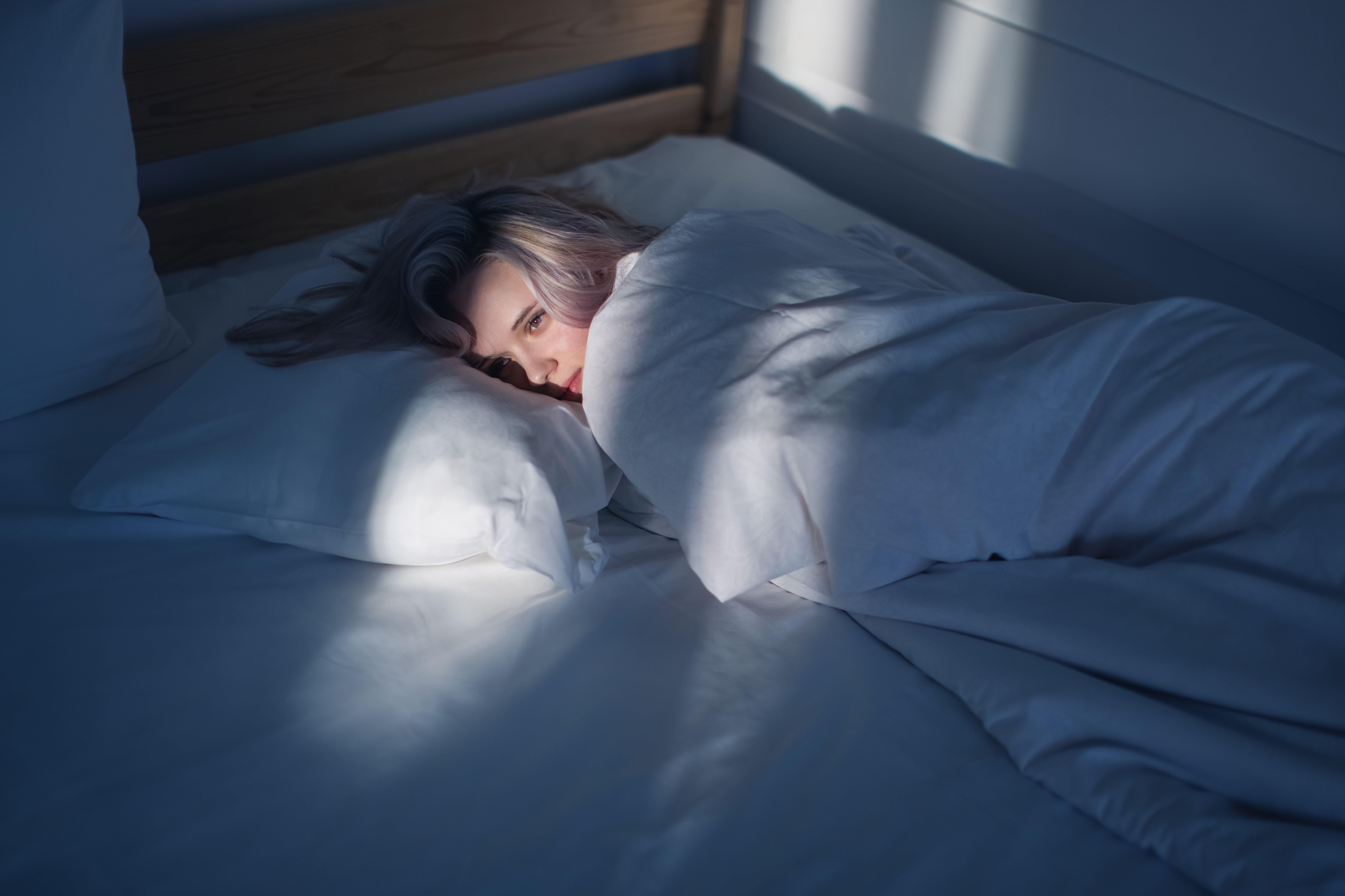 Blonde Frau liegt wach unter einer weißen Decke im Bett und schaut nachdenklich.