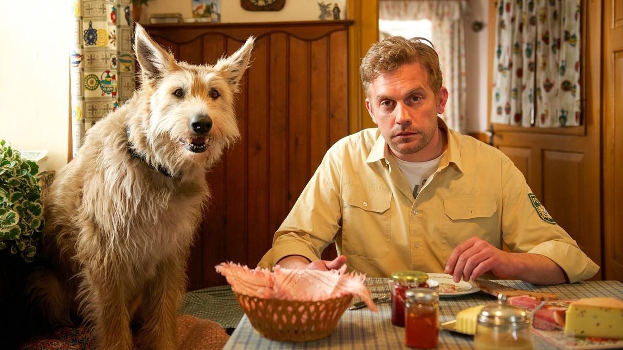 Ein Mann sitzt am Esstisch neben einem großen, zotteligen Hund in einer gemütlichen Küche.