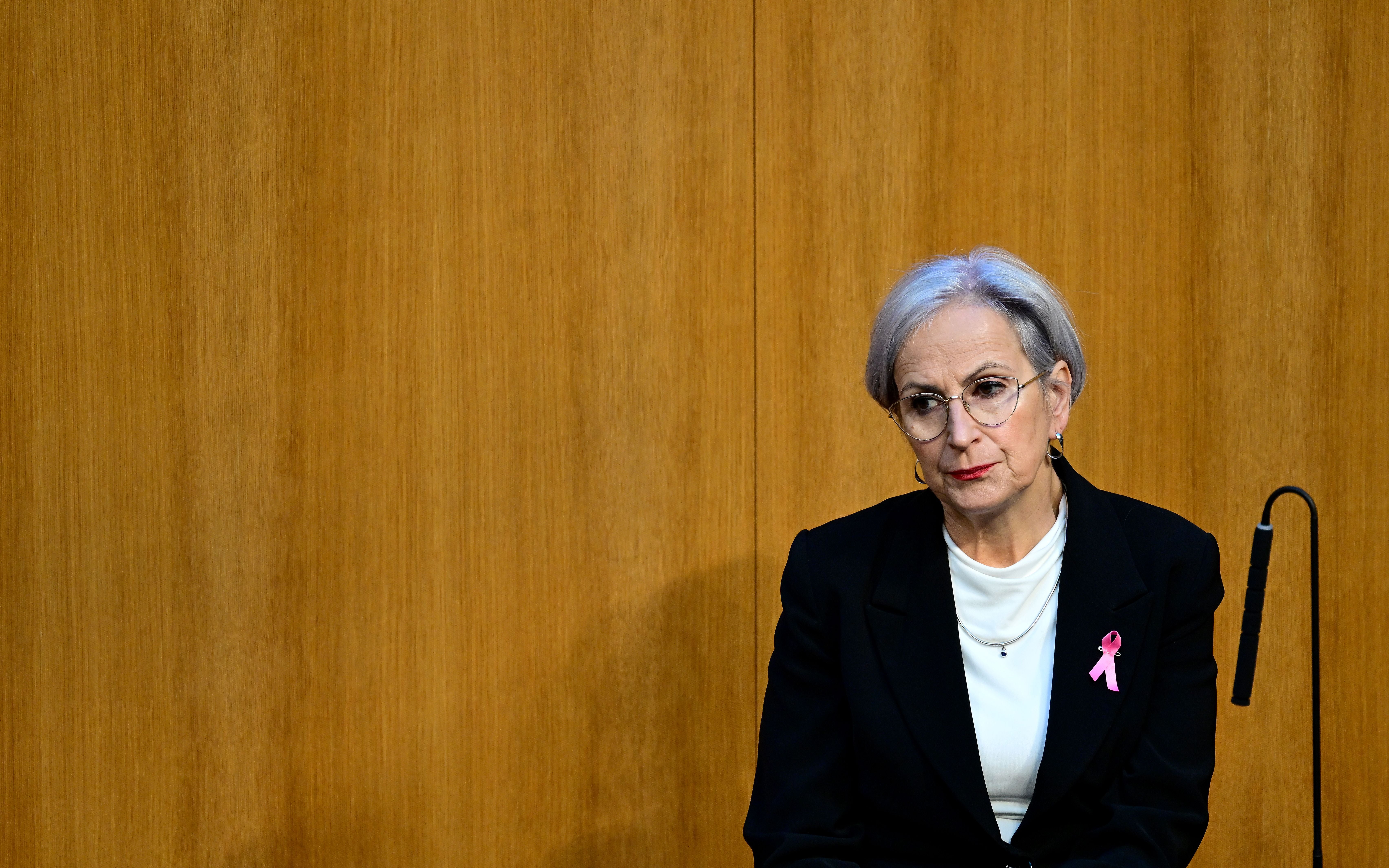 Eine Frau mit grauen Haaren und Brille sitzt vor einer Holzvertäfelung, trägt ein rosa Schleifenabzeichen.