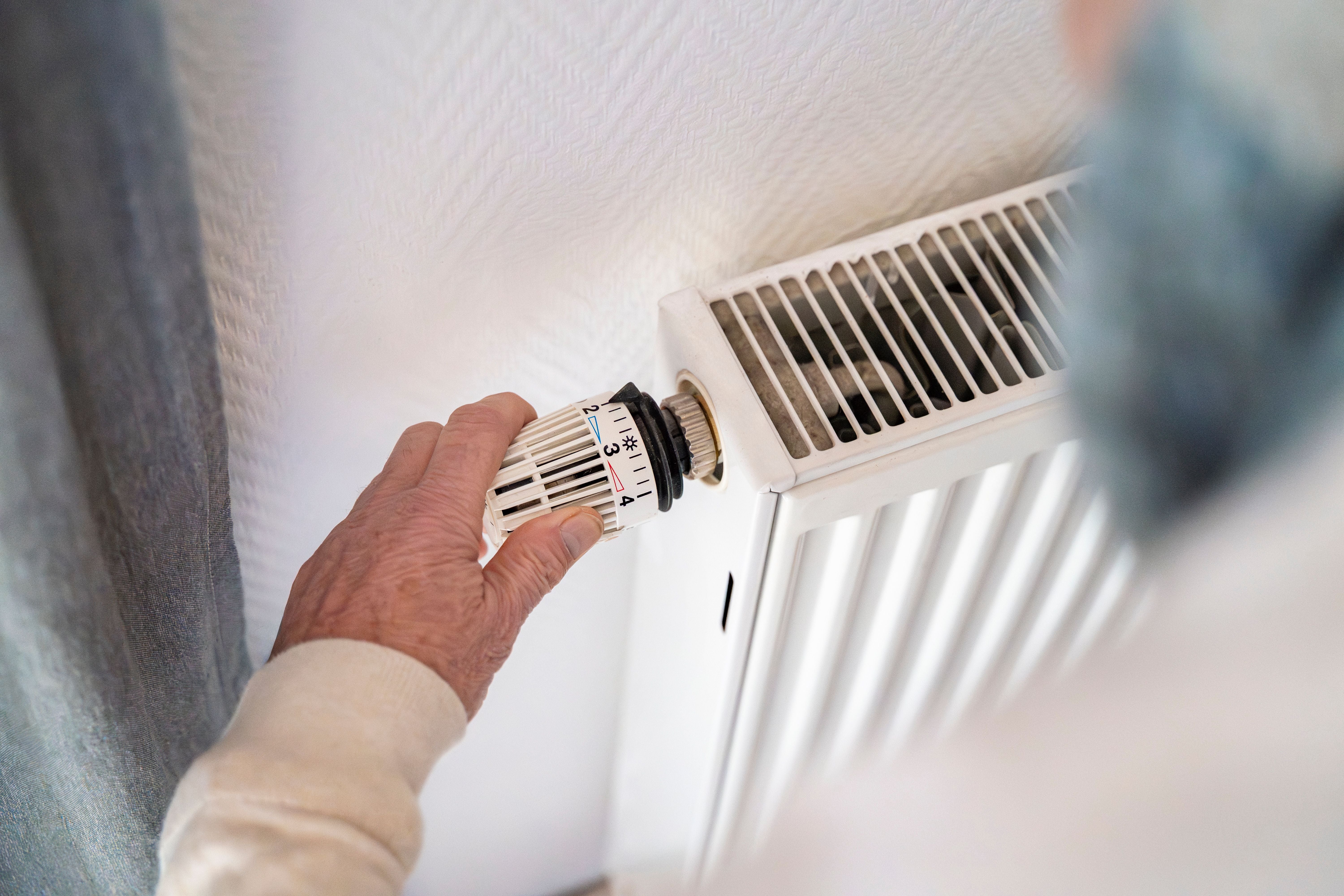 Eine Hand dreht das Thermostatventil an einer weißen Heizung in einem Raum.