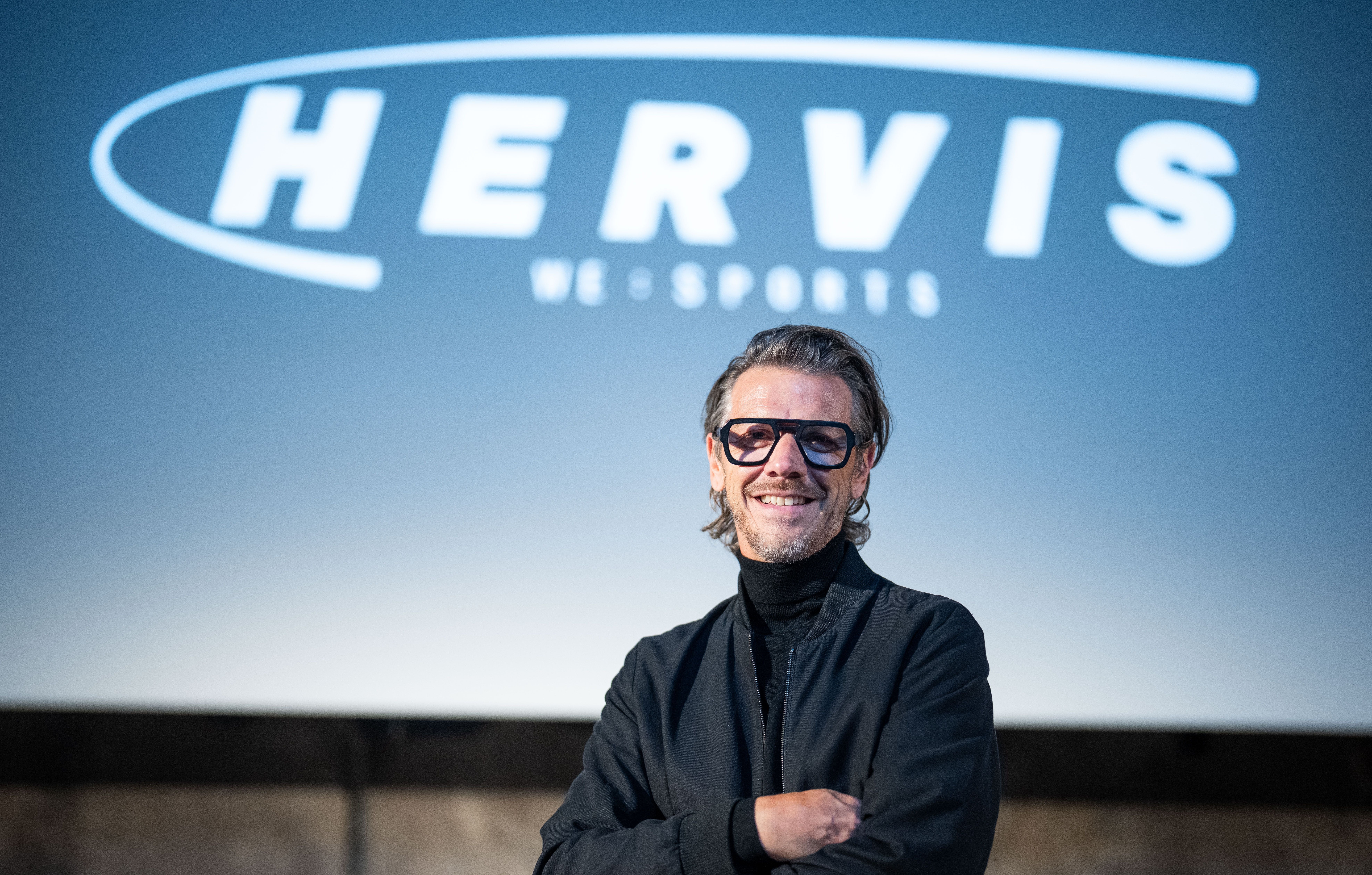 Ein Mann mit Brille steht vor einem Bildschirm mit dem Hervis-Logo und lächelt in die Kamera.