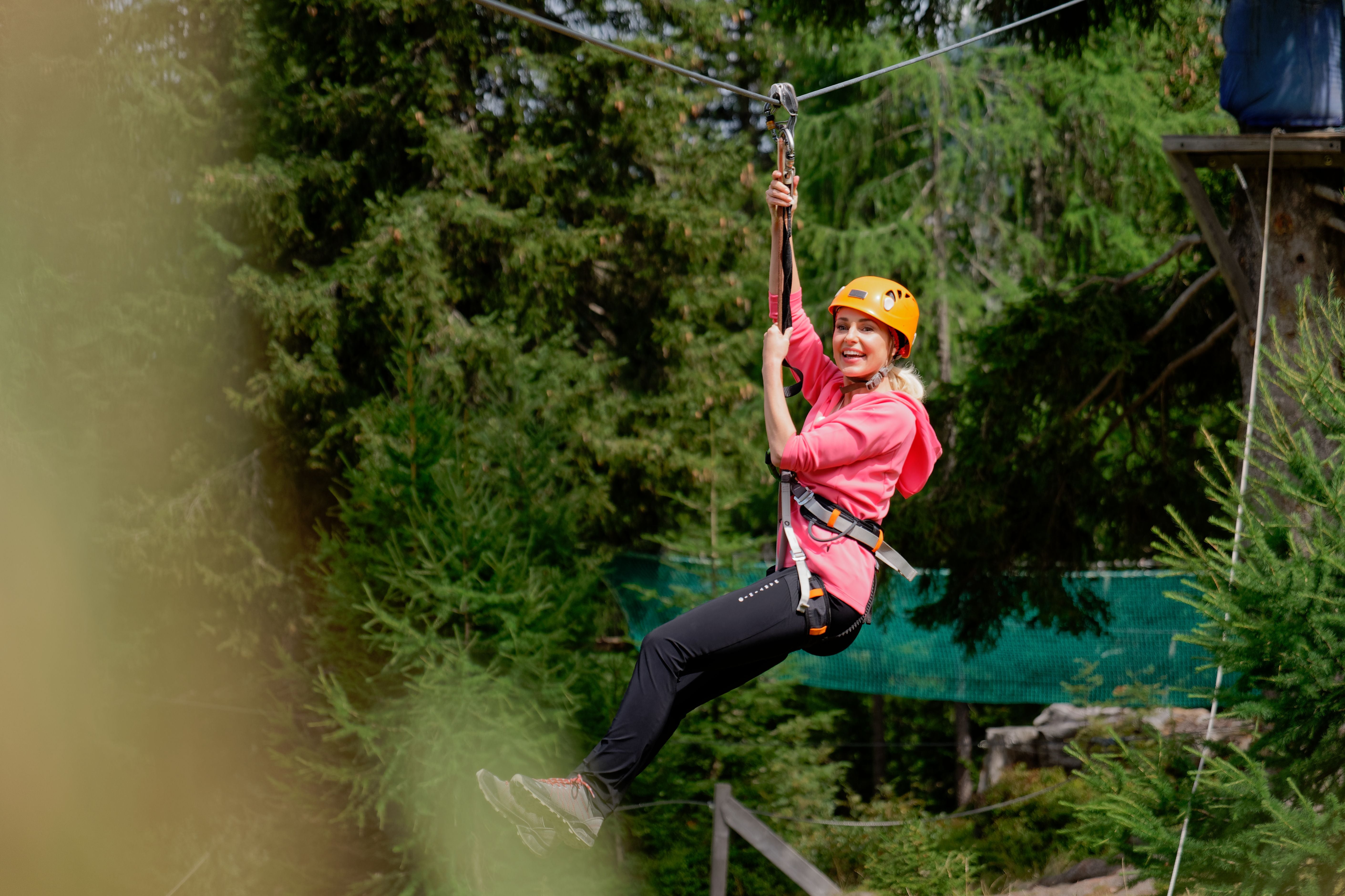 Frau mit Helm fährt lächelnd an einer Seilrutsche durch einen Wald.