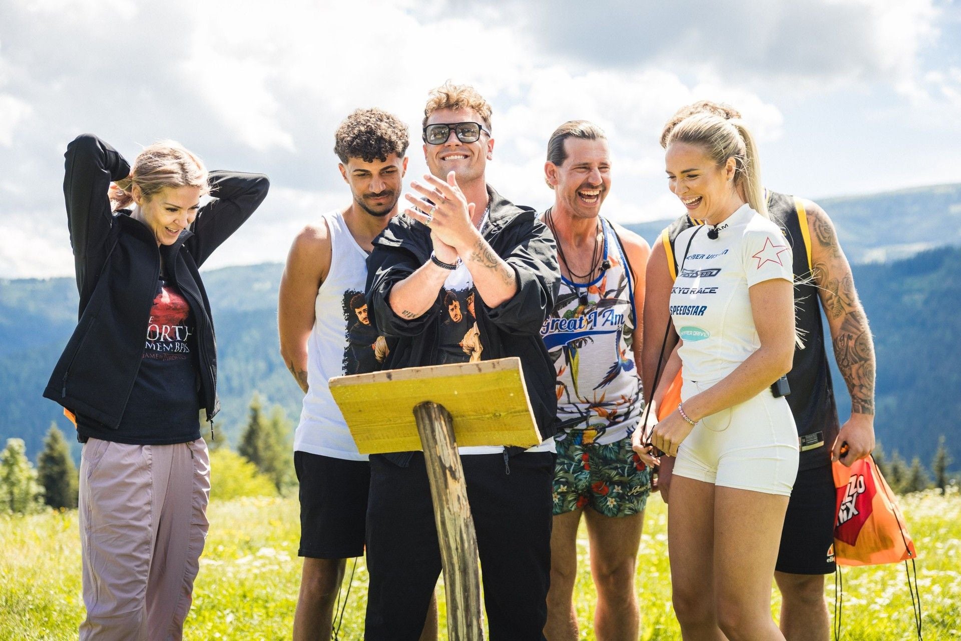 Sechs lachende Menschen stehen im Freien vor einem Holzpult auf einer Wiese in den Bergen.