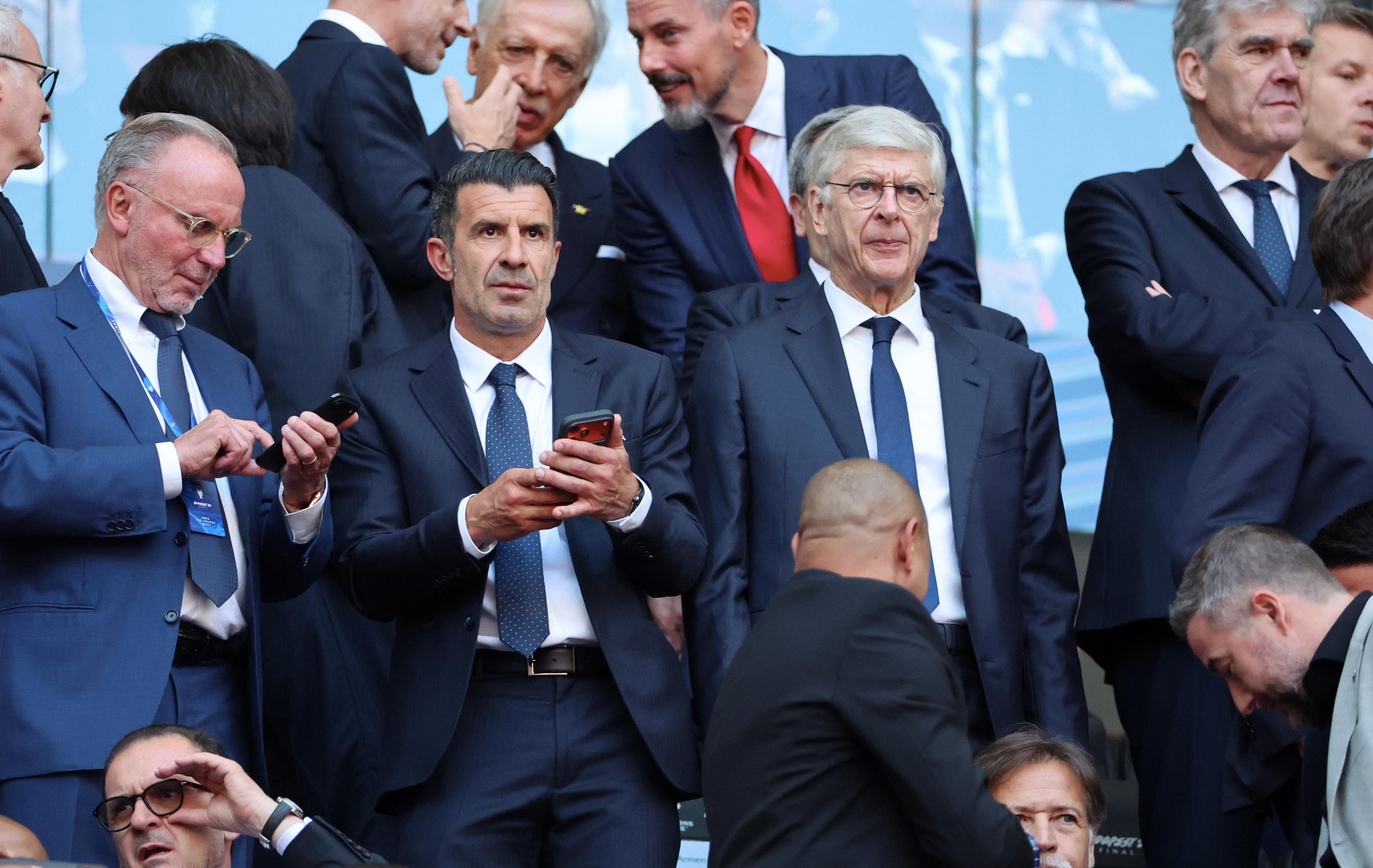 Mehrere Männer in Anzügen sitzen und stehen auf der Tribüne eines Fußballstadions.