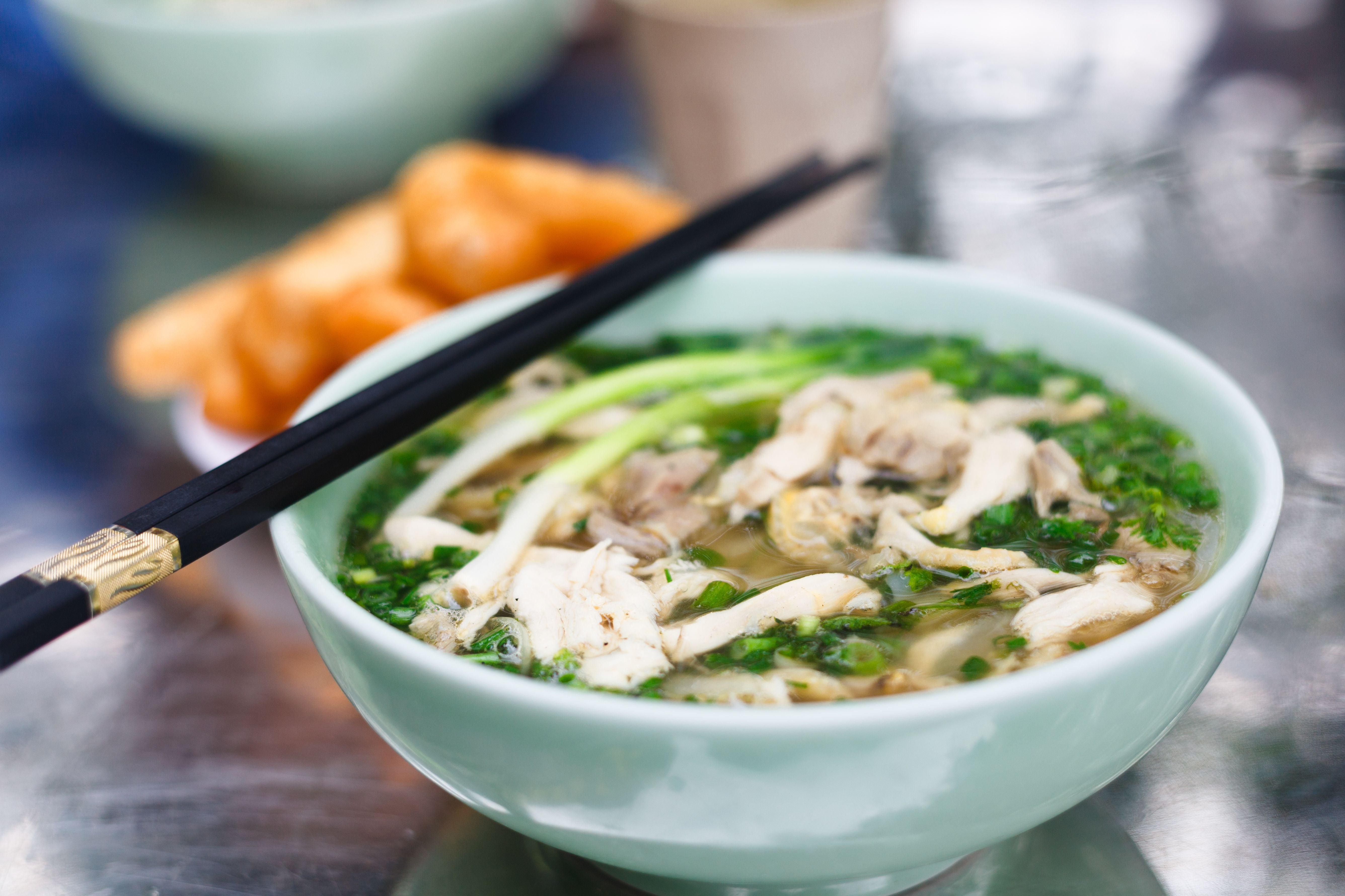 Schale mit vietnamesischer Pho-Suppe, darauf schwarze Essstäbchen, im Hintergrund Gebäck.