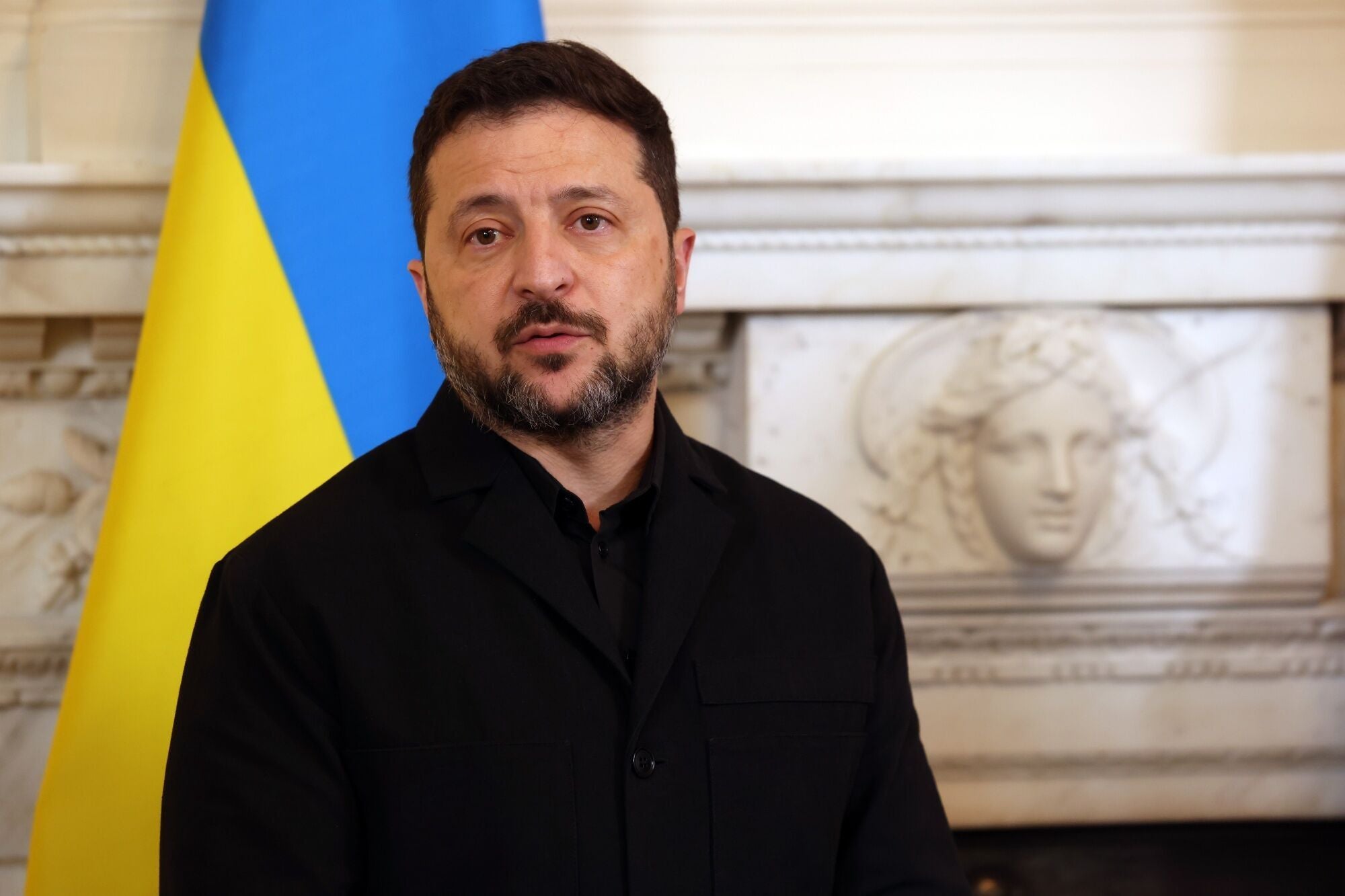 Ein Mann spricht vor einer ukrainischen Flagge und einer dekorativen Wandverzierung.