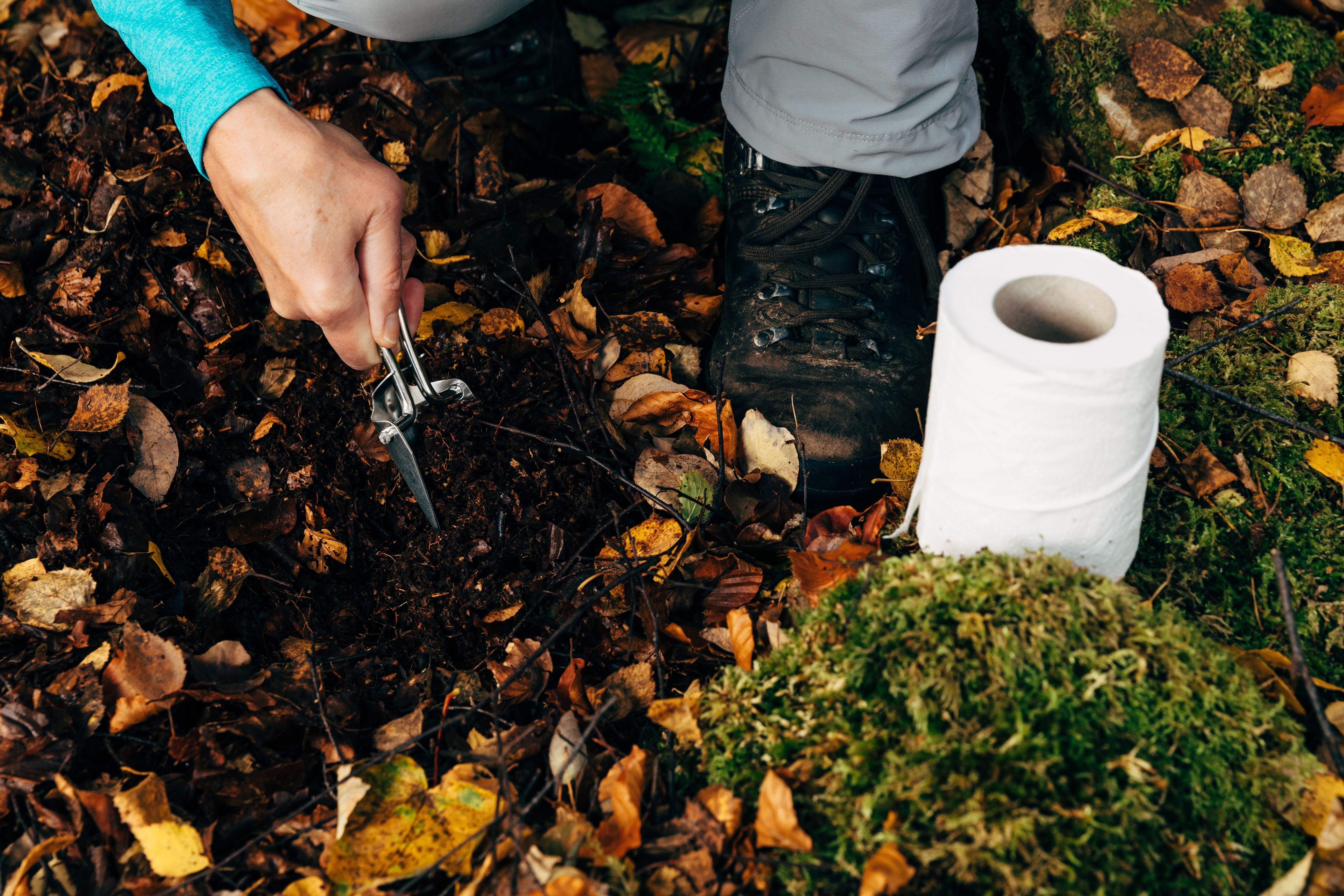 Frau gräbt mit Schaufel ein Loch im Wald, daneben liegt eine Rolle Toilettenpapier.