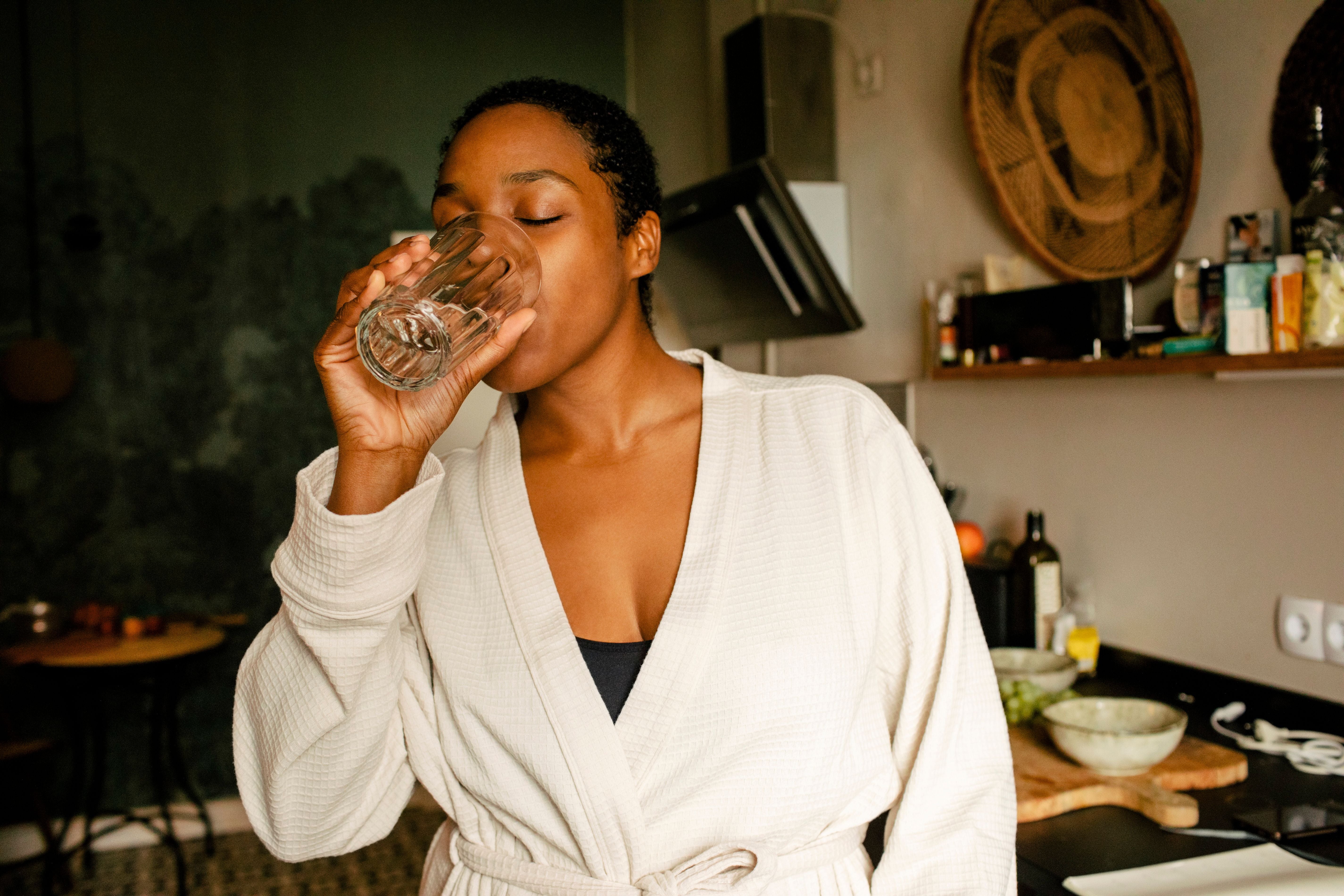 Frau im Bademantel trinkt ein Glas Wasser in einer Küche.