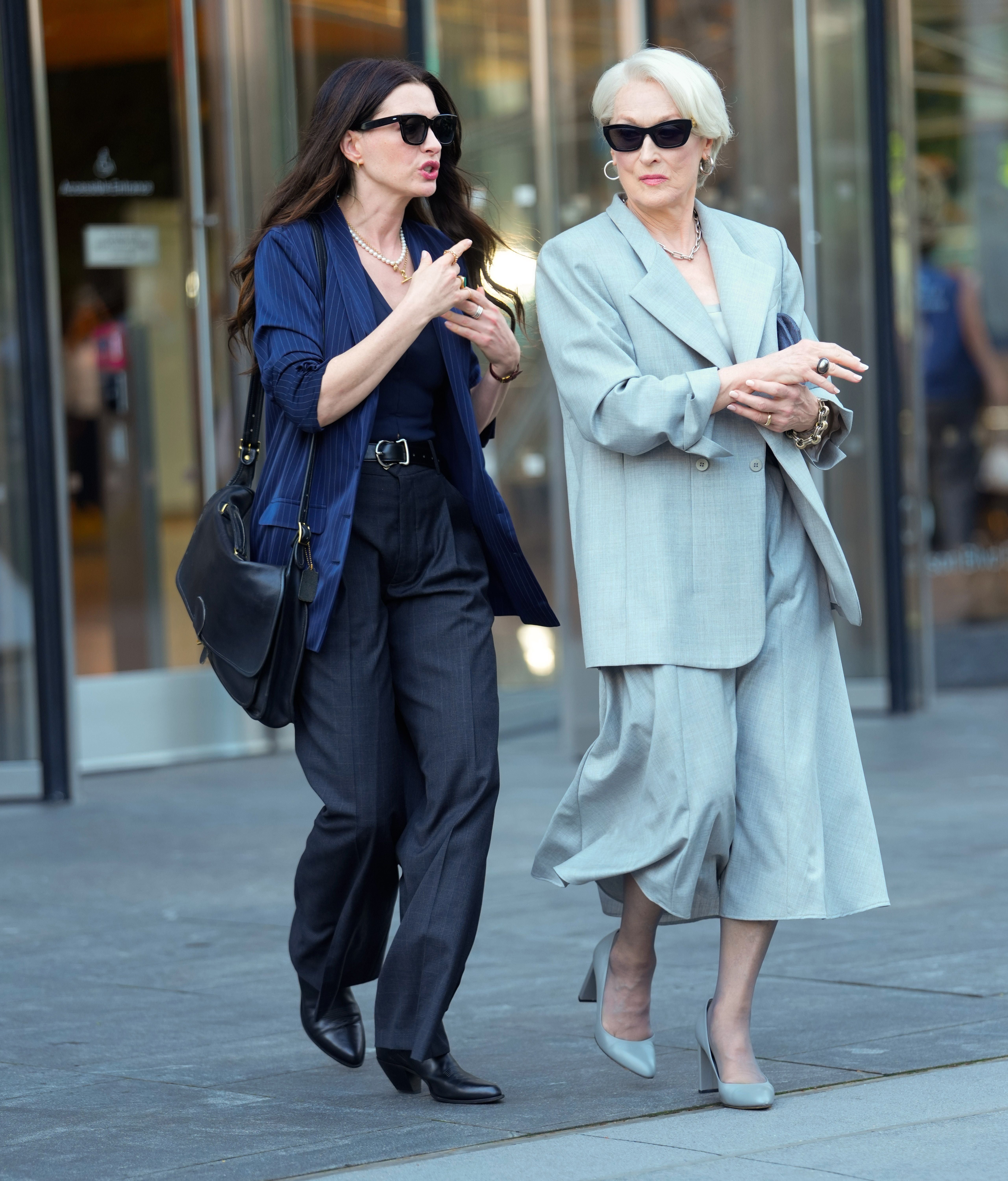 Zwei elegant gekleidete Frauen gehen nebeneinander auf einem Gehweg in einer Stadt.