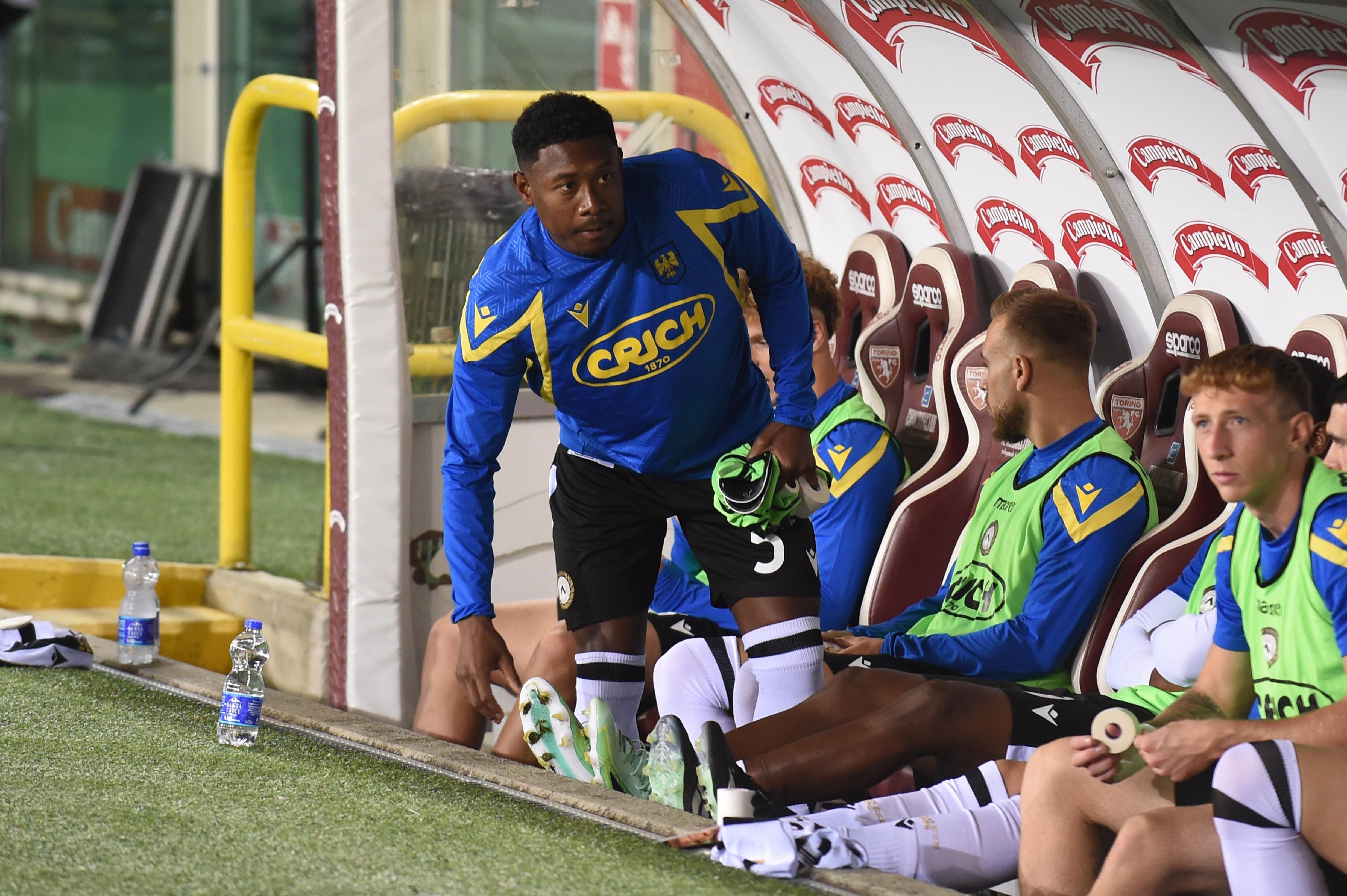 Ein Fußballspieler von Udinese steht an der Ersatzbank, umgeben von sitzenden Mitspielern.