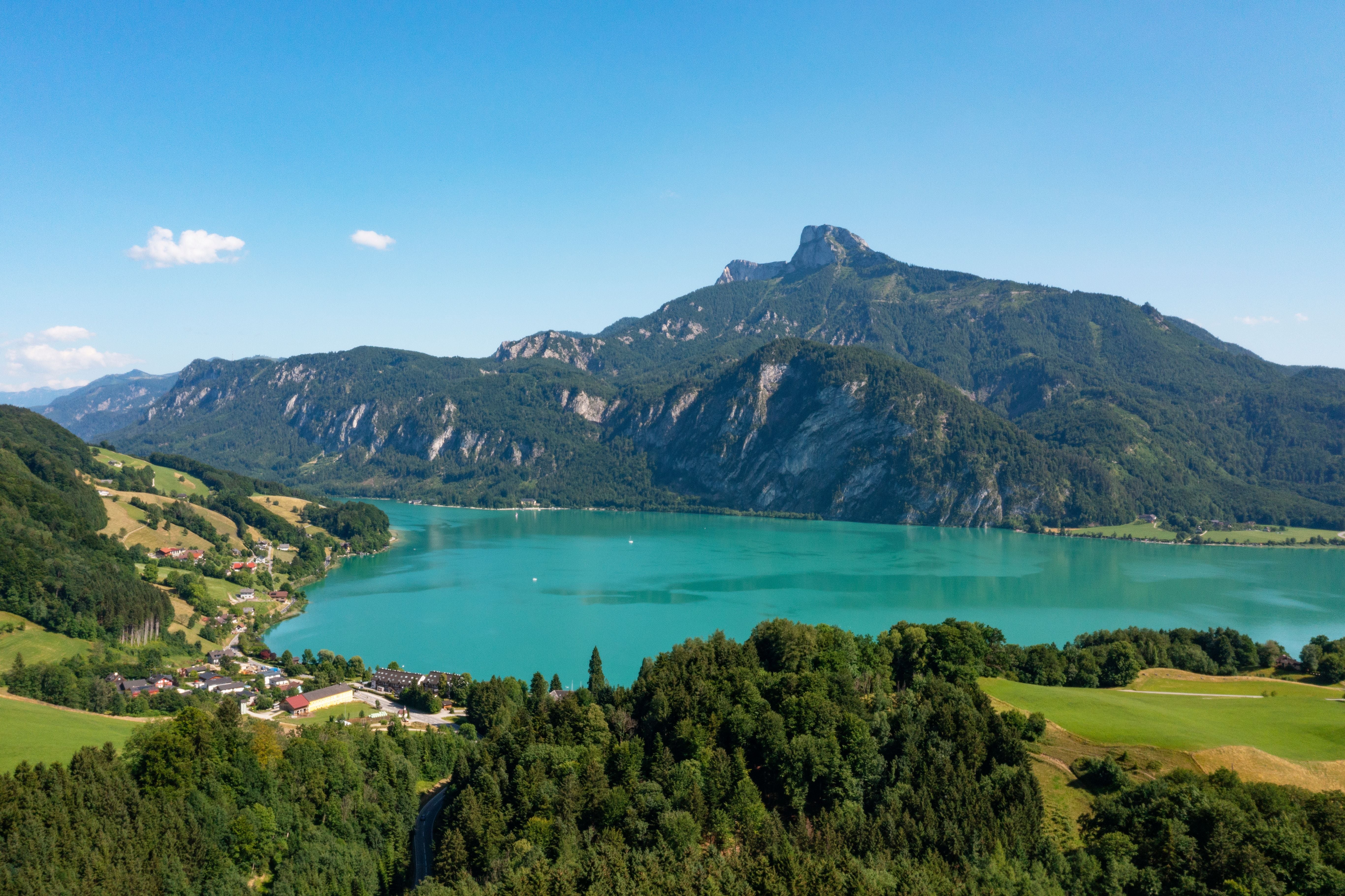 Blick auf den türkisfarbenen Mondsee mit umliegenden Wäldern und Bergen bei klarem Himmel.