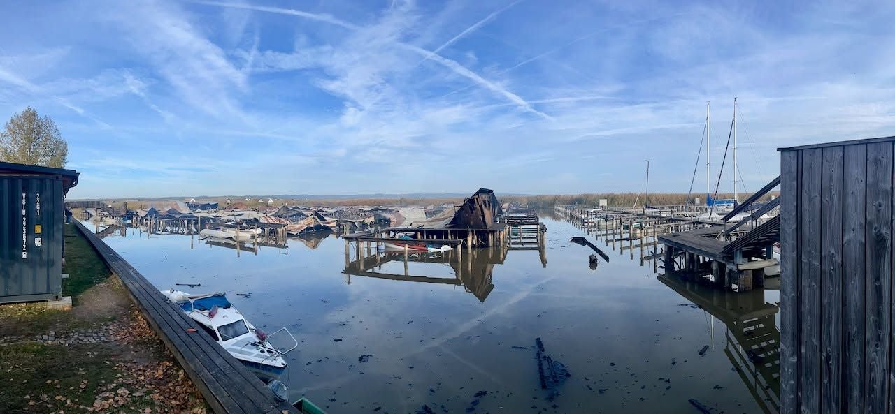Verbrannte Bootshütten in Rust nach einem Großfeuer, zerstörte Stege spiegeln sich im Wasser.