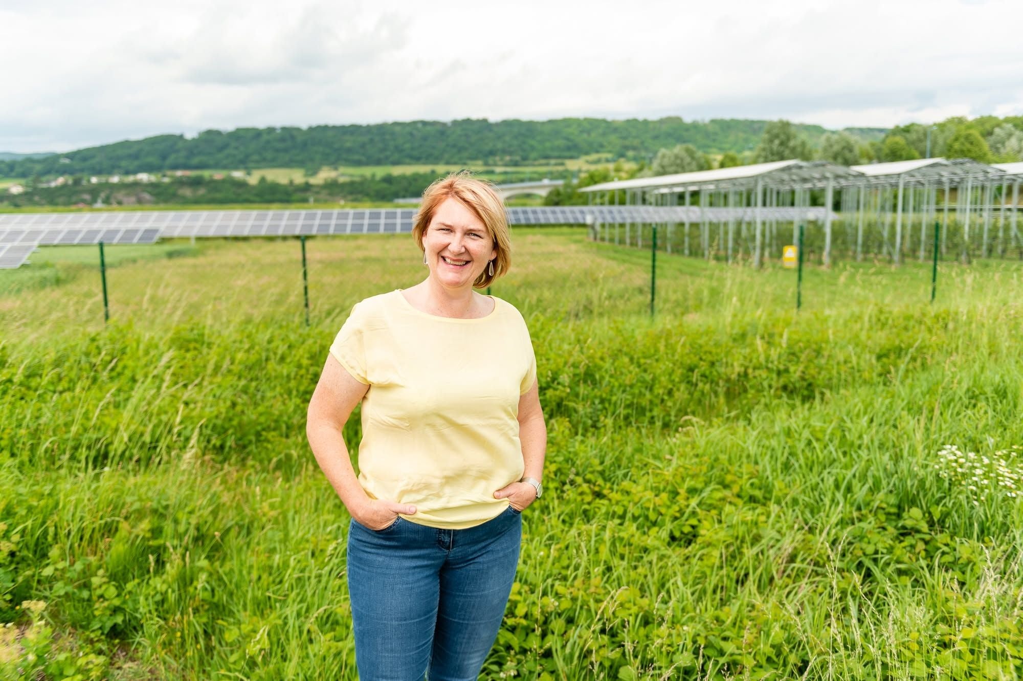 Frau mit gelbem Shirt steht lächelnd vor Solarpanels auf einer grünen Wiese.