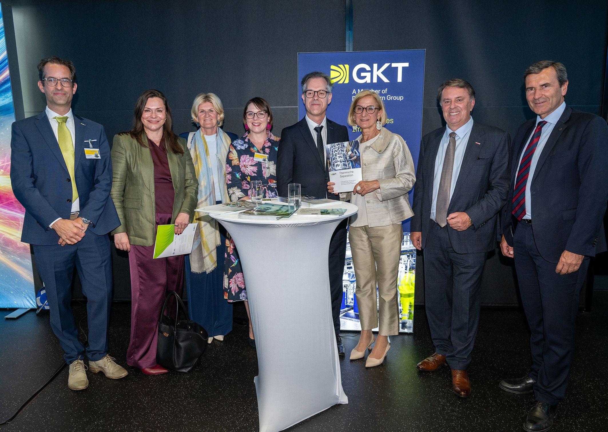 Acht Personen stehen um einen Stehtisch bei einer Veranstaltung von GKT und posieren für ein Foto.
