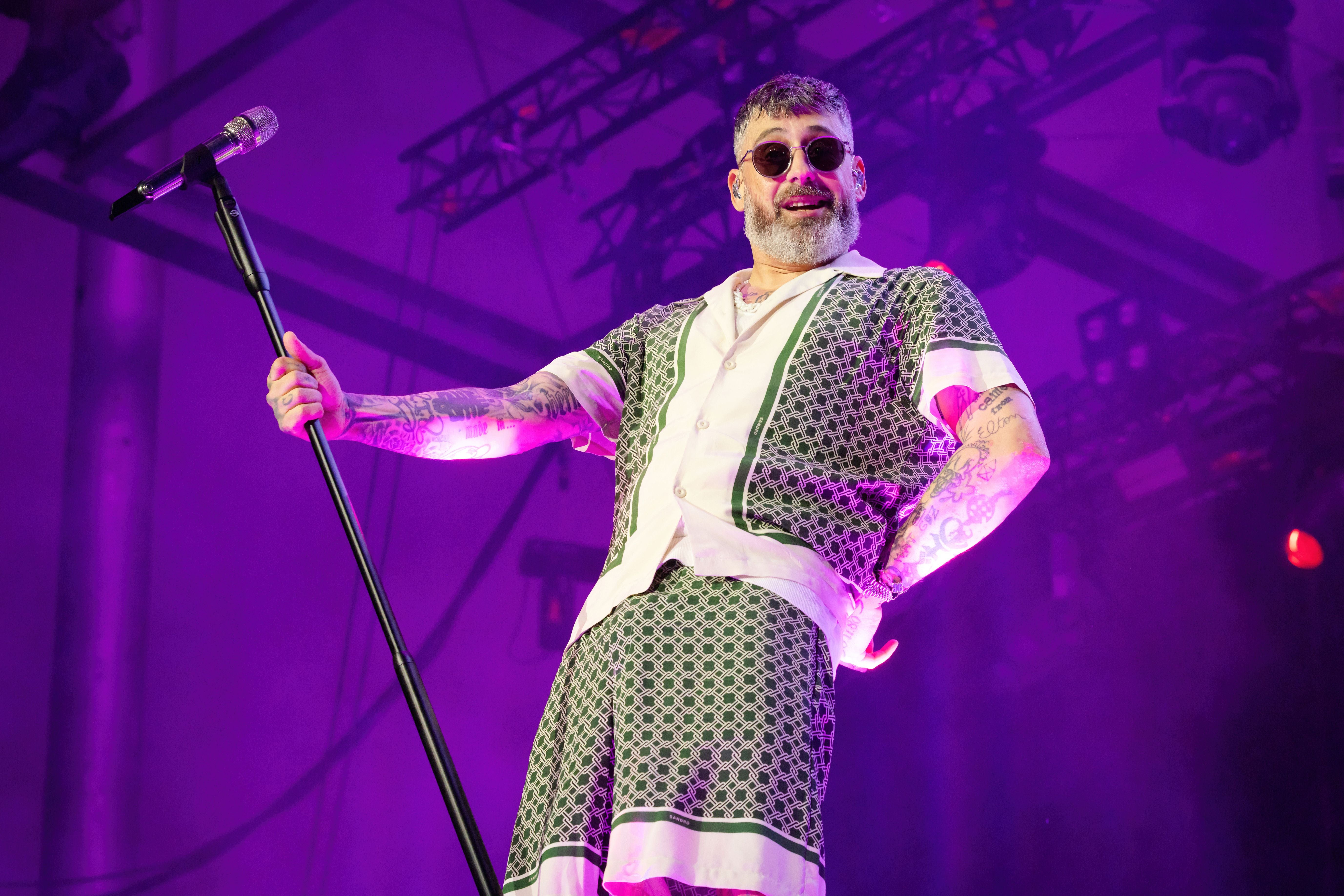 Sänger mit Sonnenbrille und gemustertem Outfit singt auf einer Bühne mit lila Beleuchtung.