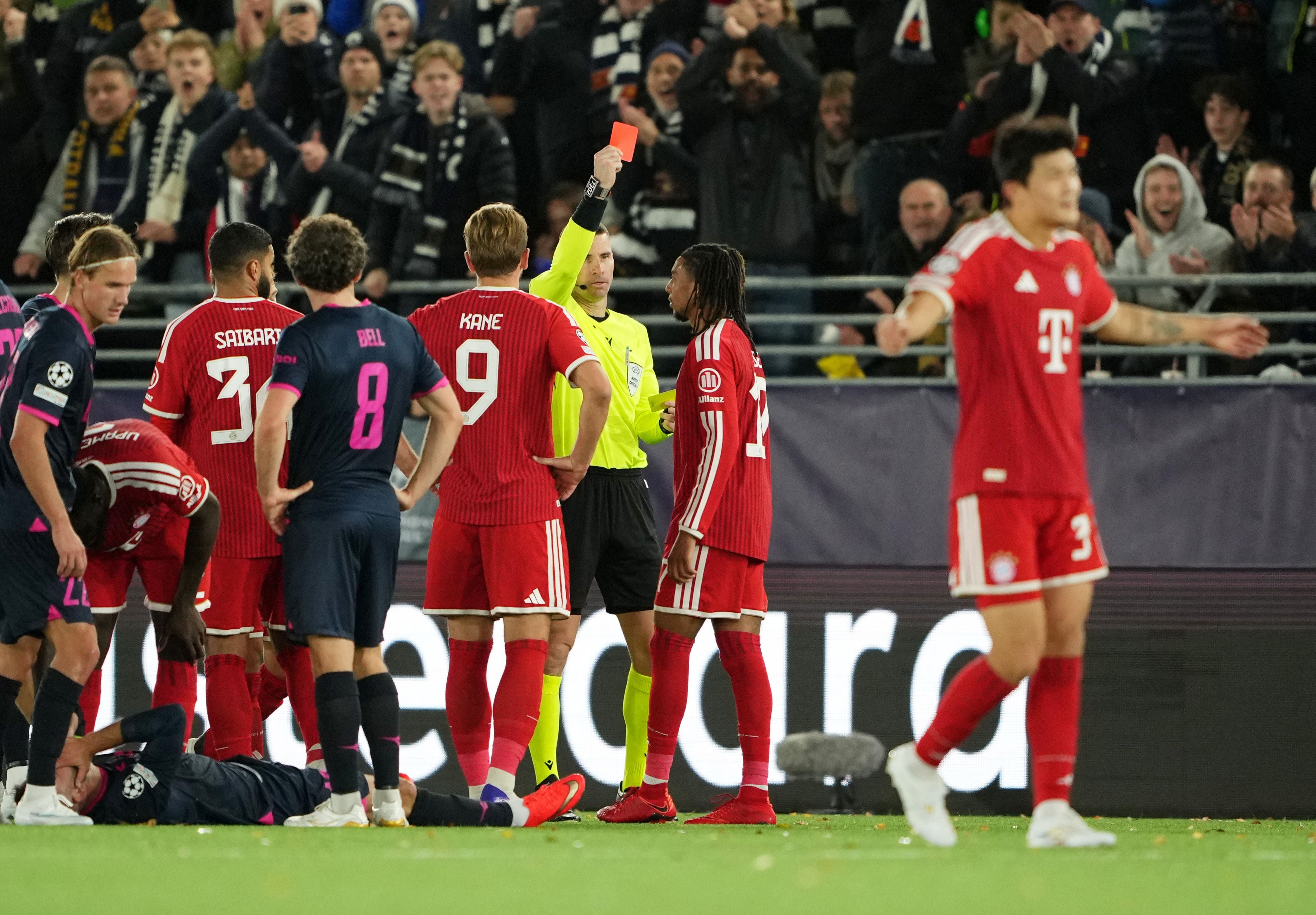 Ein Schiedsrichter zeigt einem Bayern-Spieler im Fußballspiel eine rote Karte, Spieler und Fans reagieren.