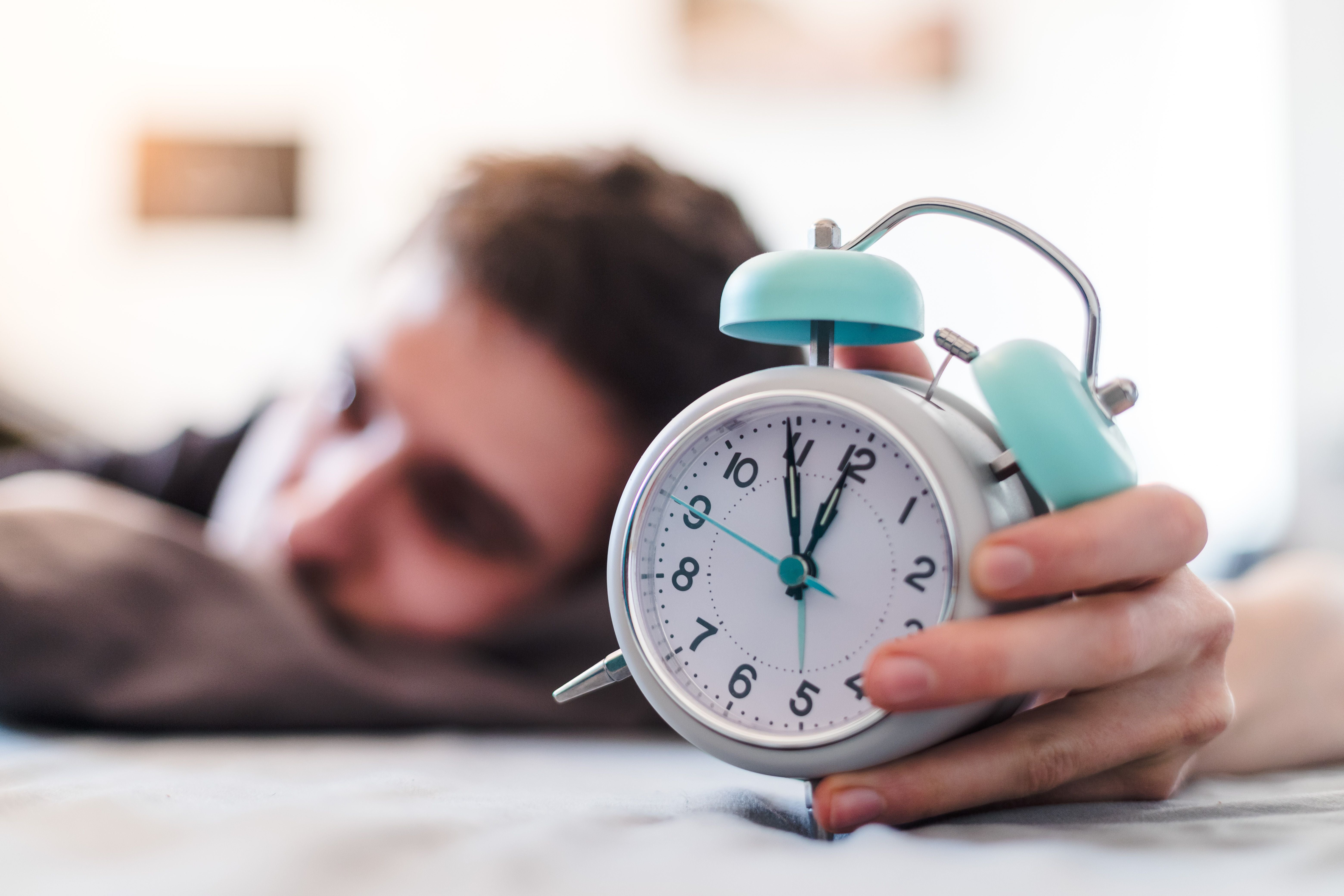 Junger Mann liegt im Bett und hält einen weißen Wecker in der Hand.