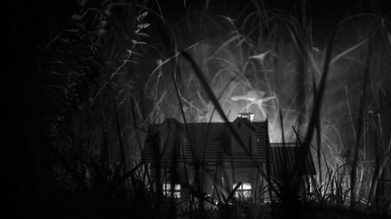 Kleines beleuchtetes Haus inmitten von hohem Gras bei Nacht, stimmungsvolles Schwarzweißfoto.