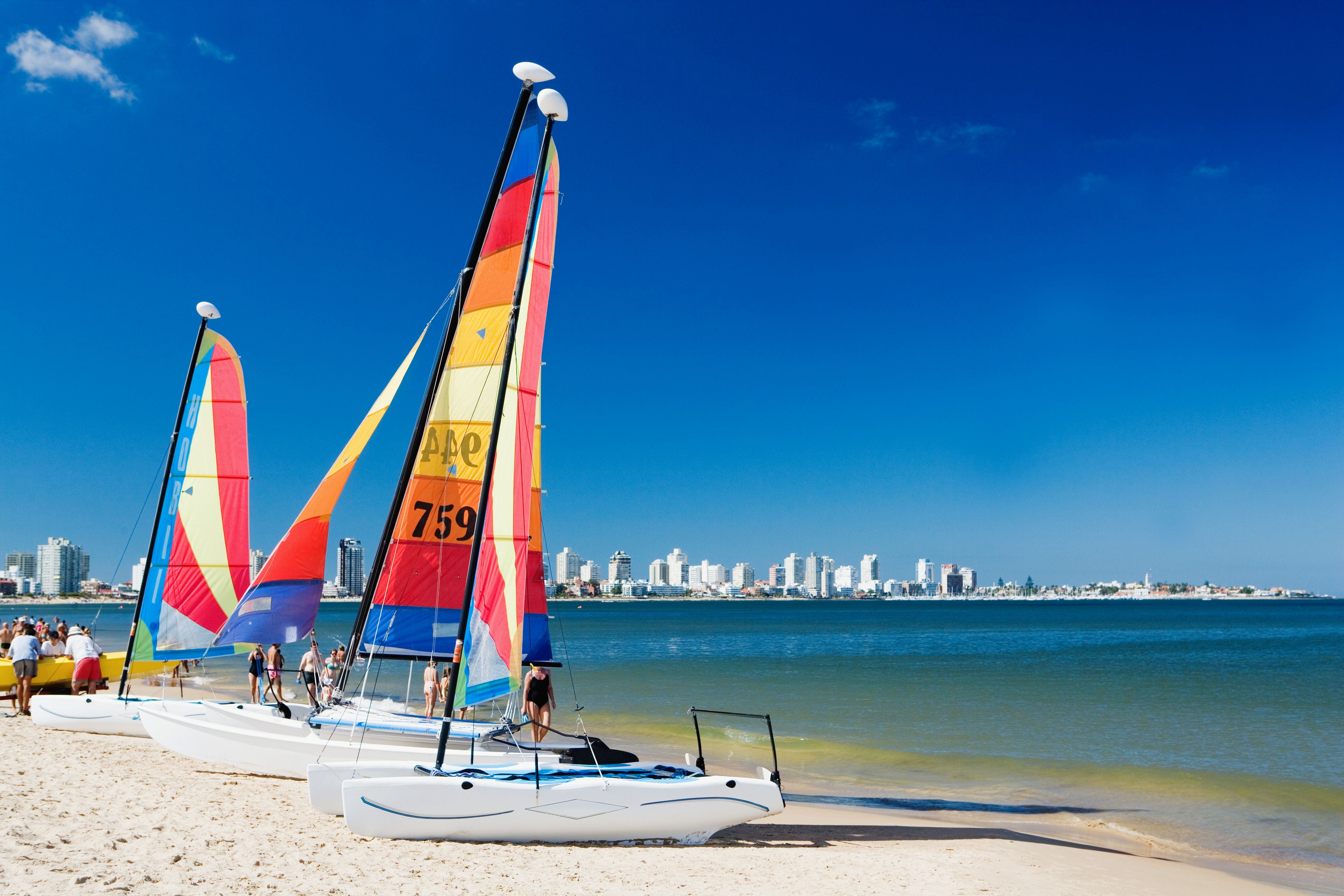 Drei bunte Segelboote stehen am Sandstrand, im Hintergrund ist eine Skyline am Wasser zu sehen.
