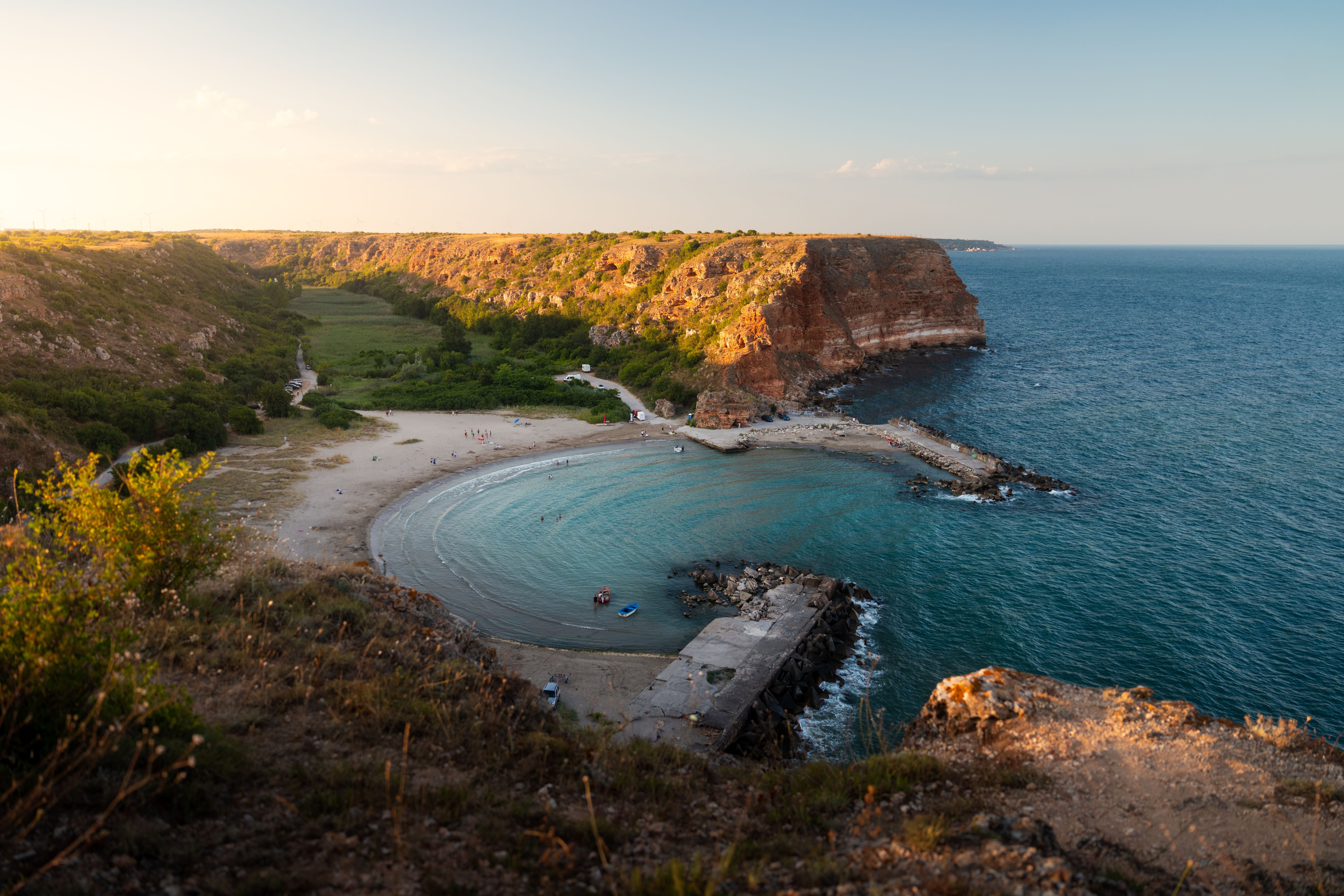 Bolata Beach mit sandiger Bucht, ruhigem türkisfarbenem Meer und roten Küstenklippen bei Sonnenuntergang.