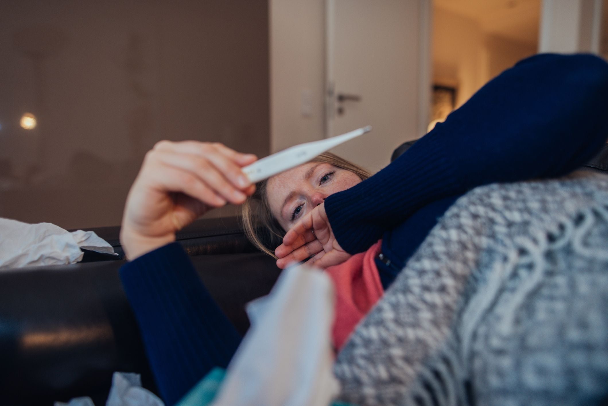 Kranke Frau liegt auf dem Sofa und schaut auf ein Fieberthermometer.
