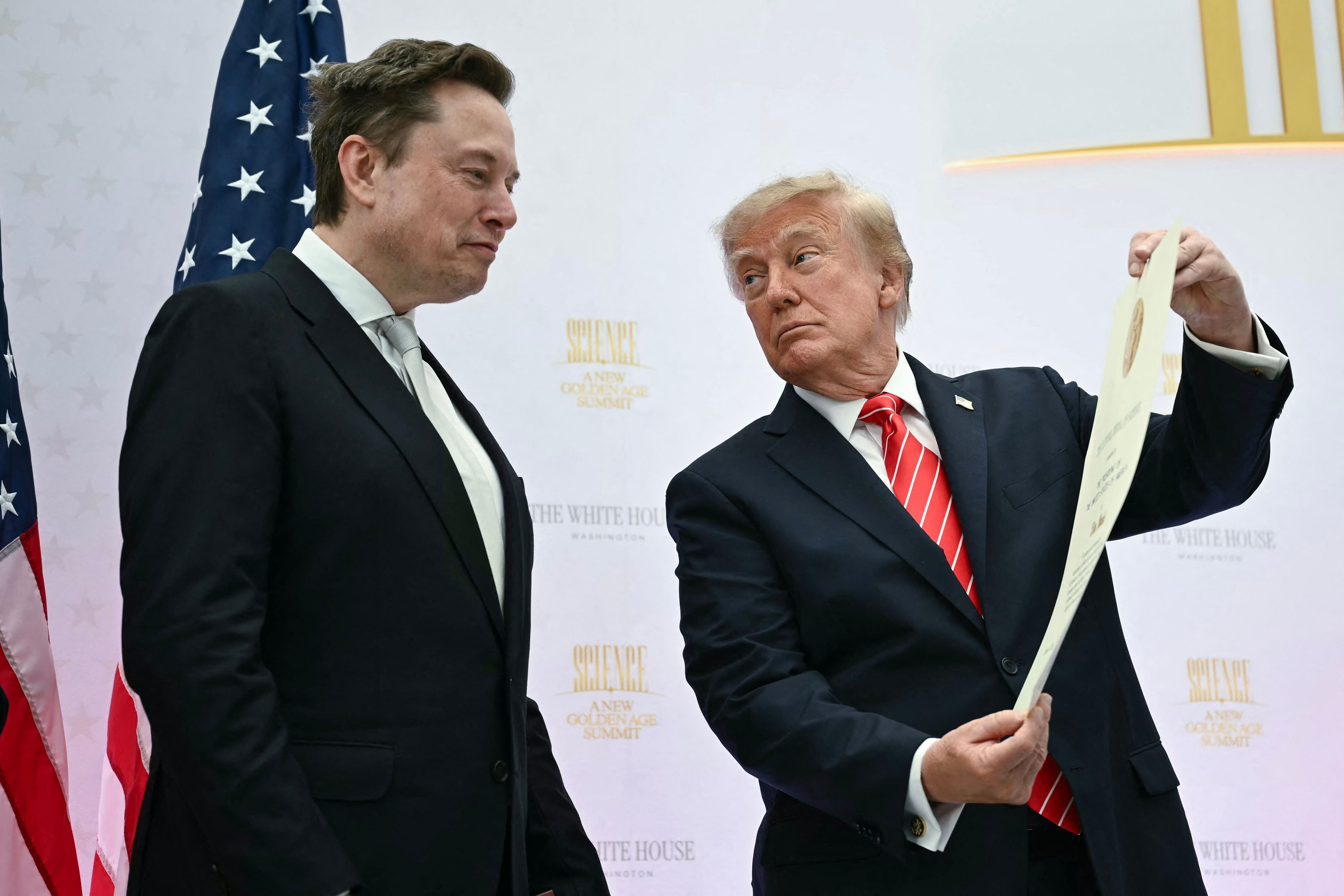 Ein Mann überreicht einem anderen Mann eine Urkunde bei einer Zeremonie, US-Flaggen im Hintergrund.