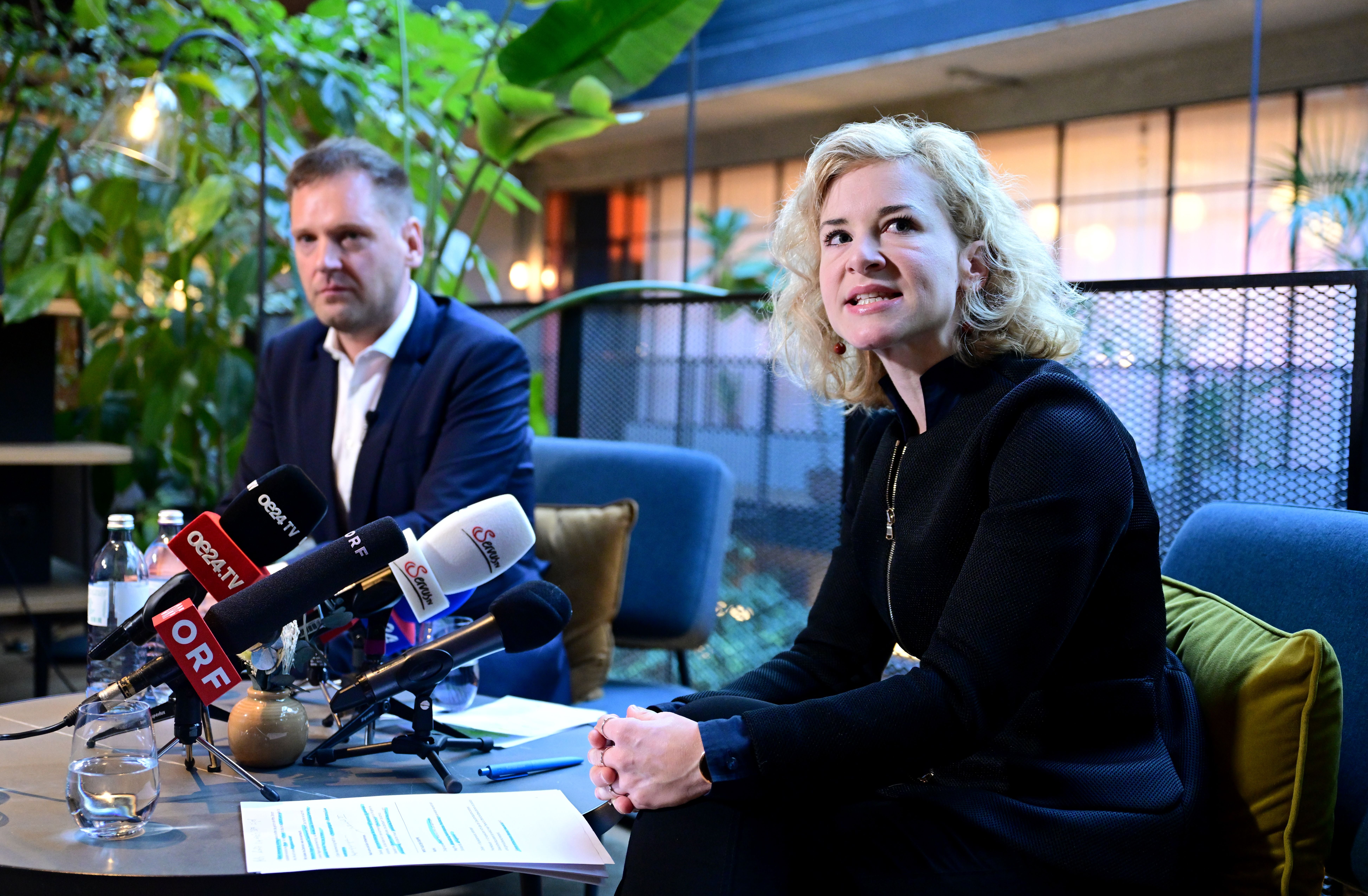 Zwei Personen sitzen bei einer Pressekonferenz mit mehreren Mikrofonen vor sich in einem modernen Raum.