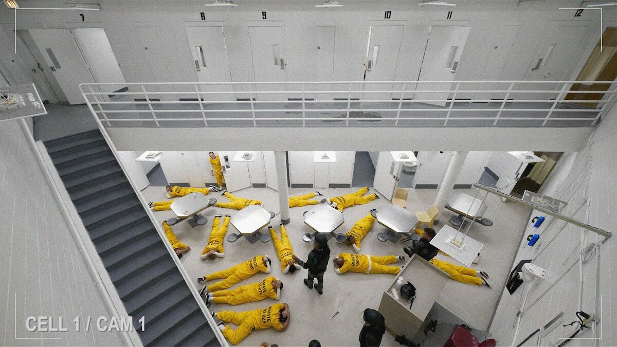 Mehrere Häftlinge in gelben Overalls liegen in einem modernen Gefängnisflur auf dem Boden.